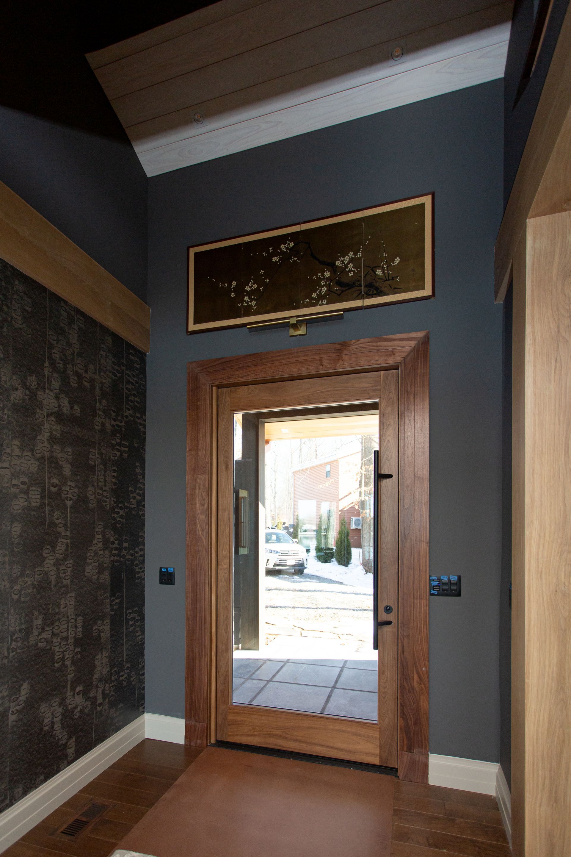 Wooden door framed in wood, set in a dark gray wall, leading outside. A piece of art hangs above the door.