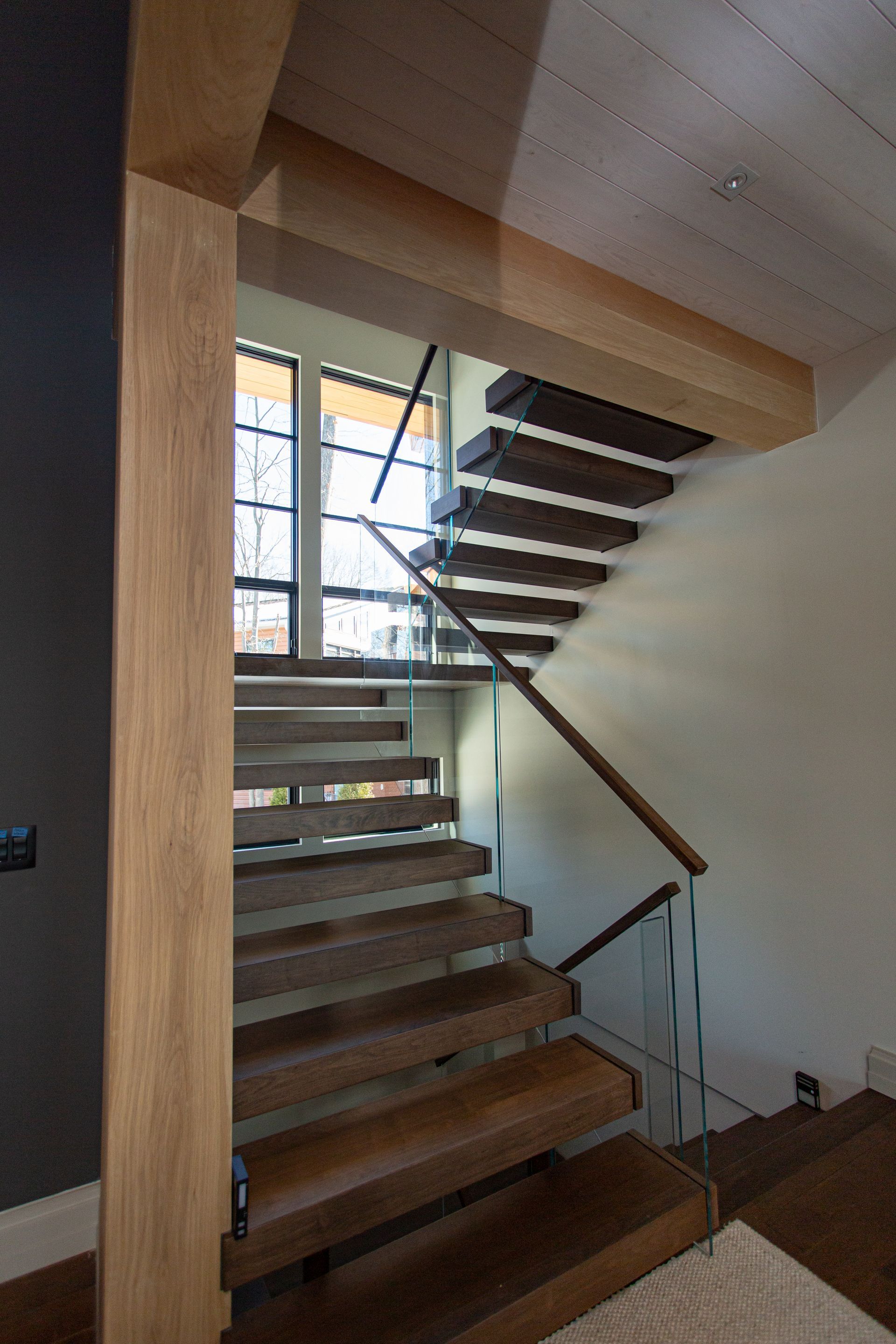 Wooden floating staircase with glass railing. Sunlight streams through a window.