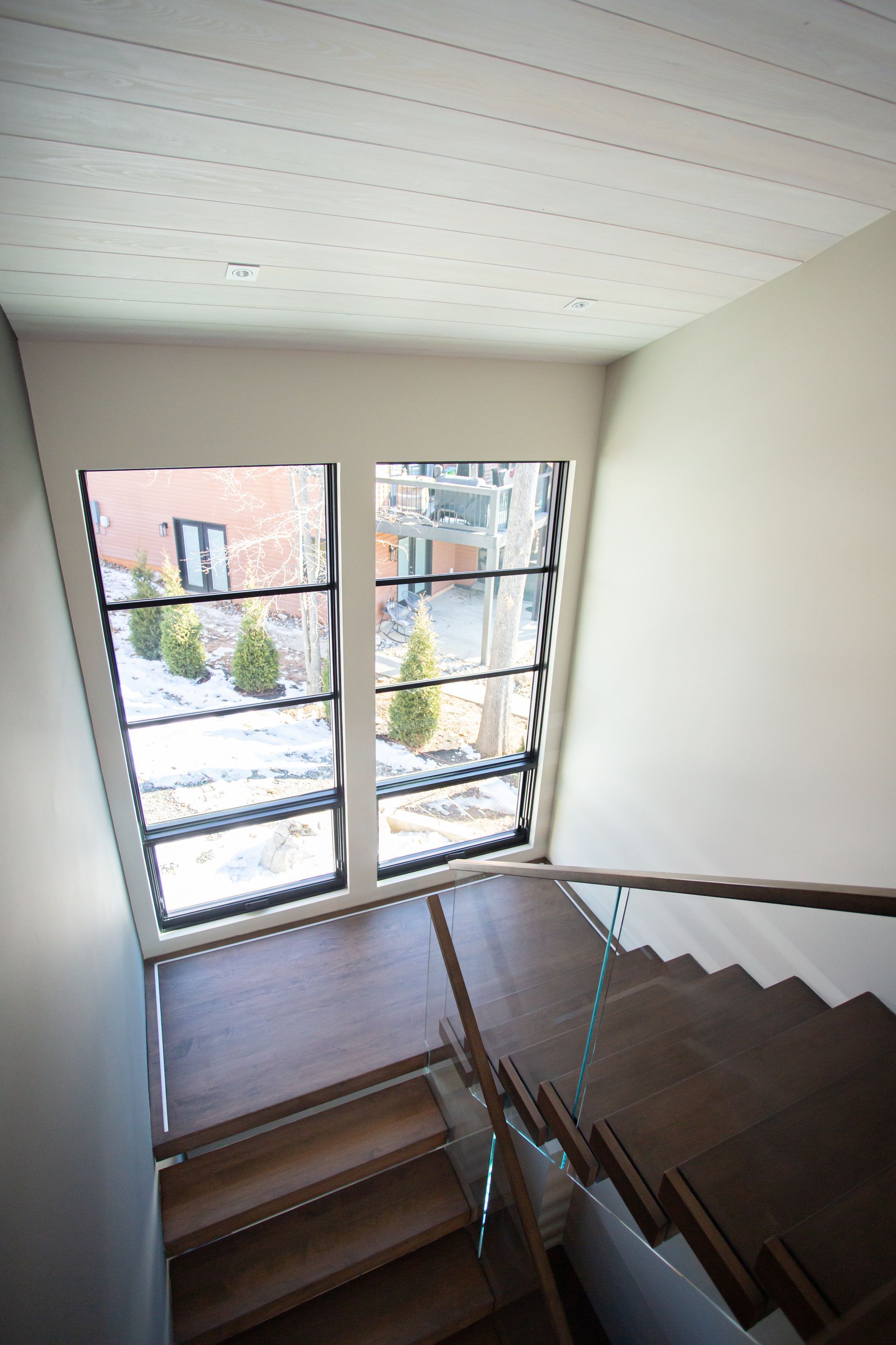 Stairwell with wooden steps, glass railing, and large angled windows overlooking a snowy outdoor scene.