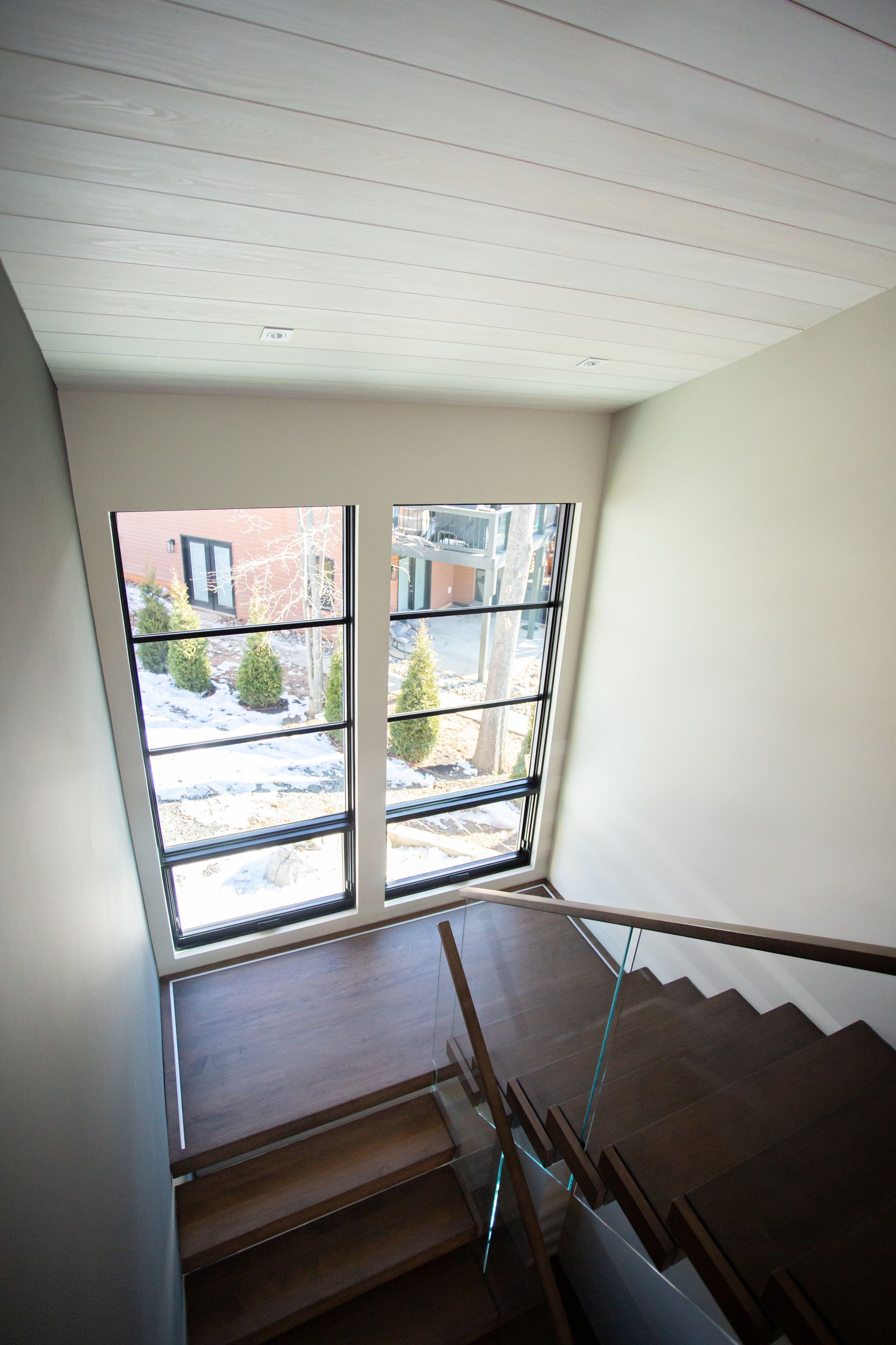 Staircase with wooden steps and glass railing leading down. Large windows show a snowy outdoor scene.