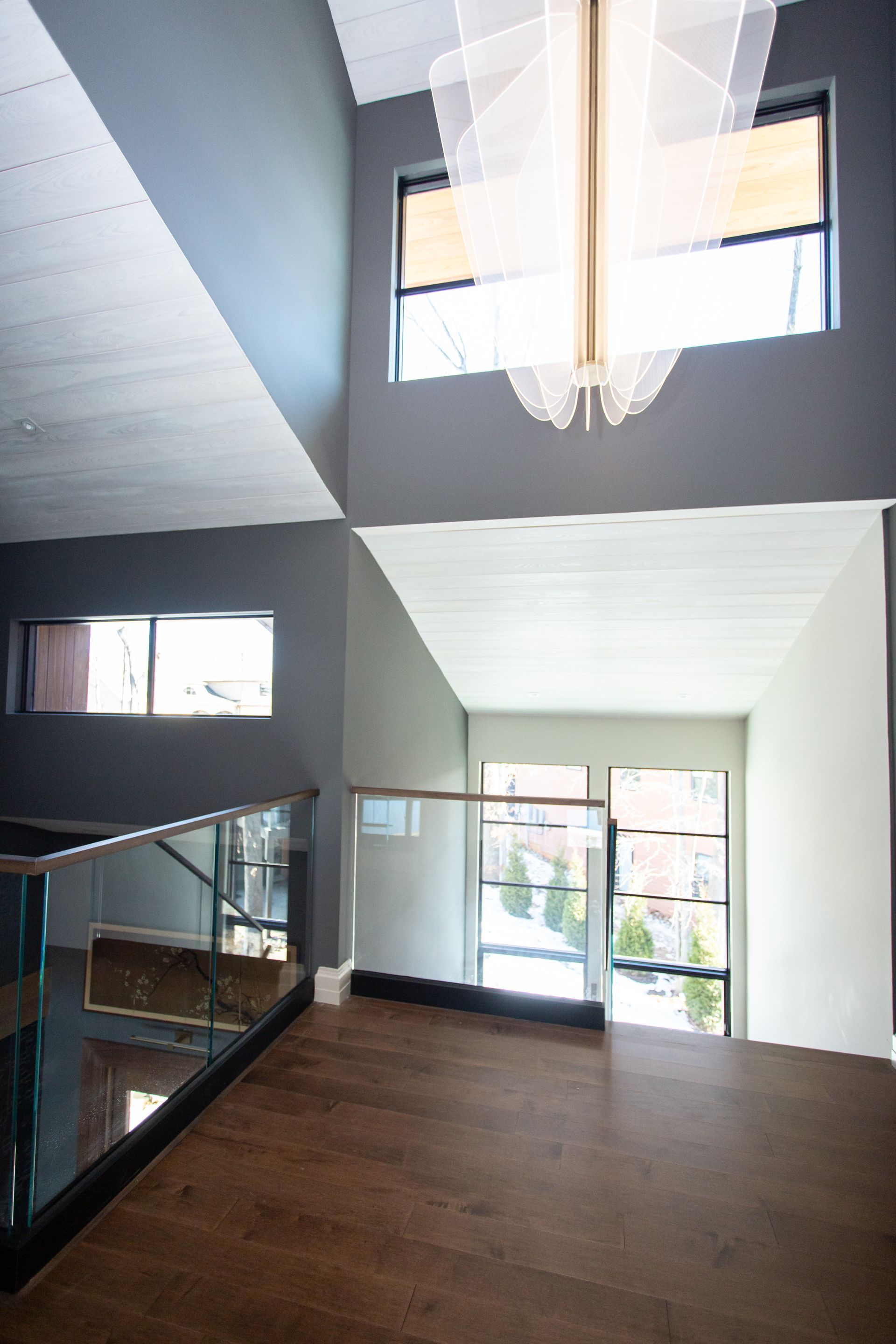 Interior view: modern home with wooden floor, glass railing, gray and white walls, windows, and large chandelier.