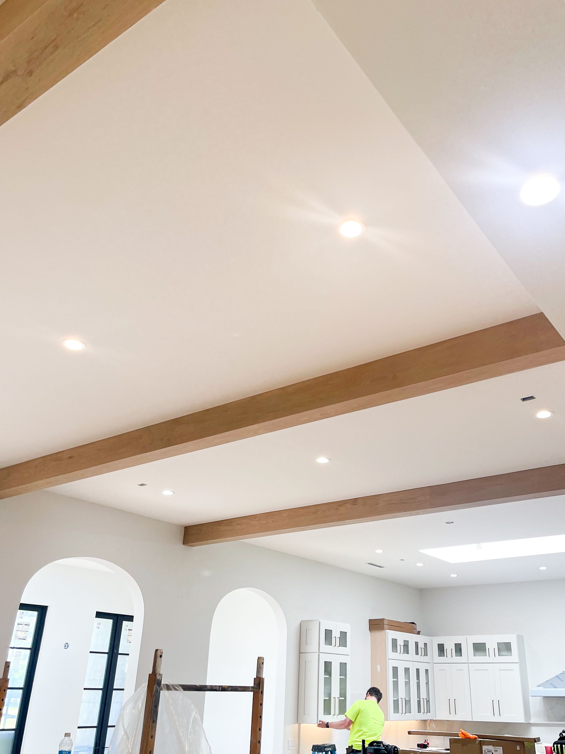 Interior view of a kitchen with light-colored ceiling, wooden beams, and arched doorways. A person in a yellow vest is working.