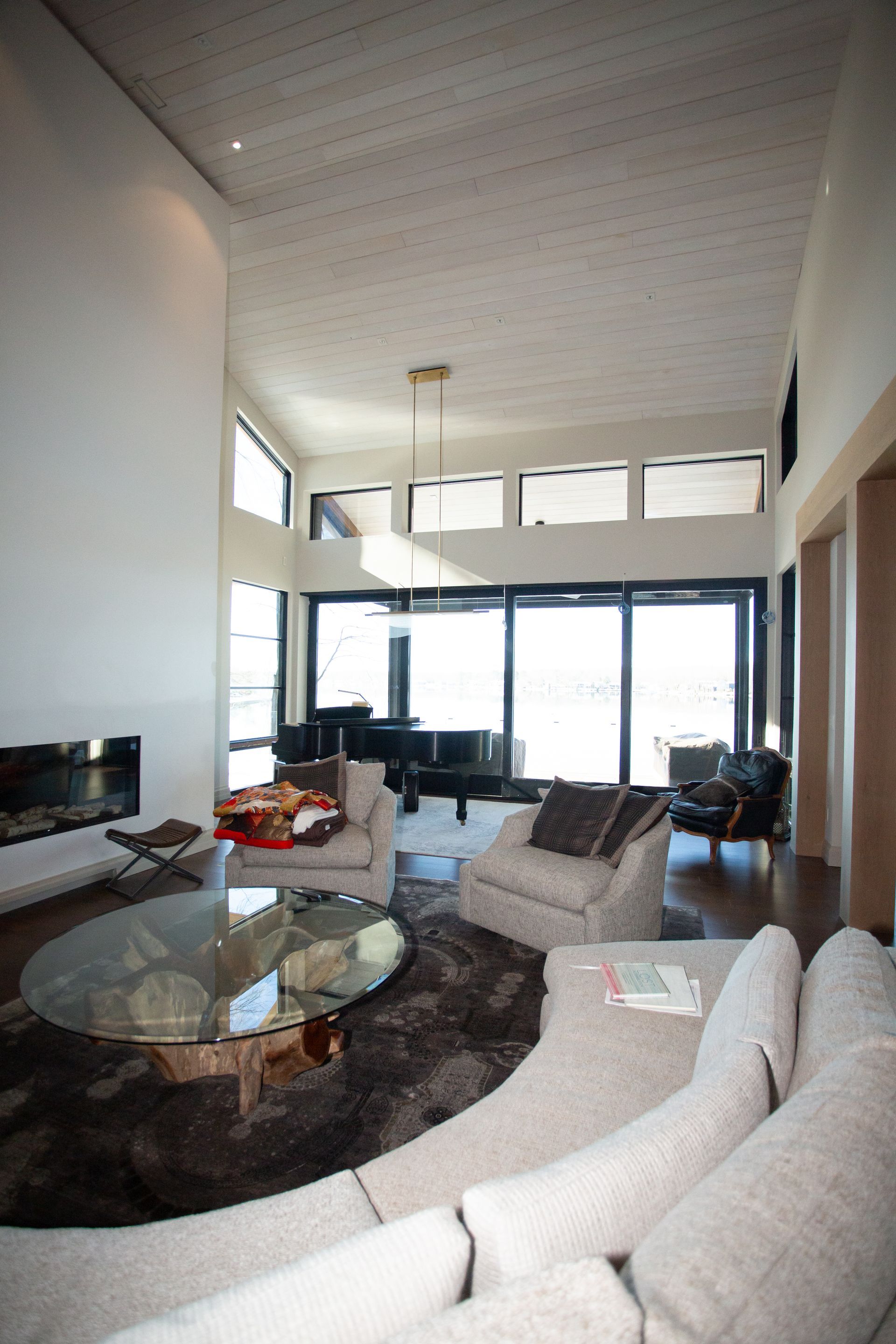 Spacious modern living room with fireplace, grand piano, and large windows overlooking outdoors. Neutral tones.