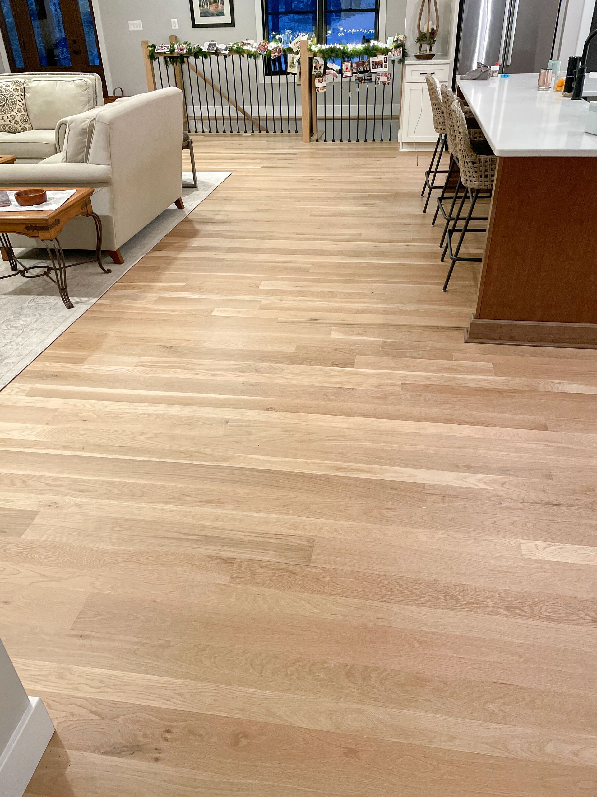Light-colored hardwood floors in a well-lit living space with kitchen island and stairs in the background.