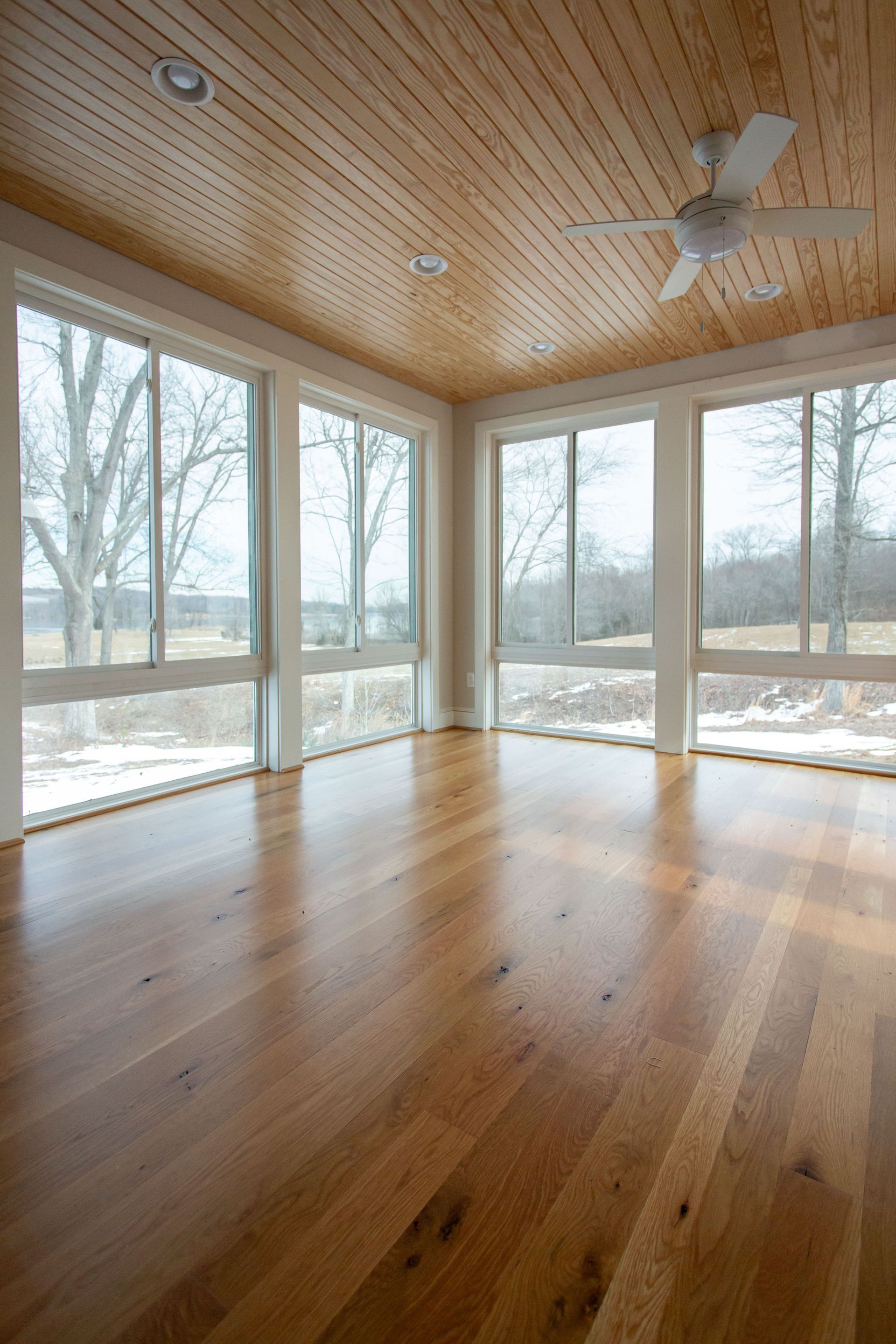 Empty sunroom with wood floors and ceiling, large windows, and a ceiling fan.