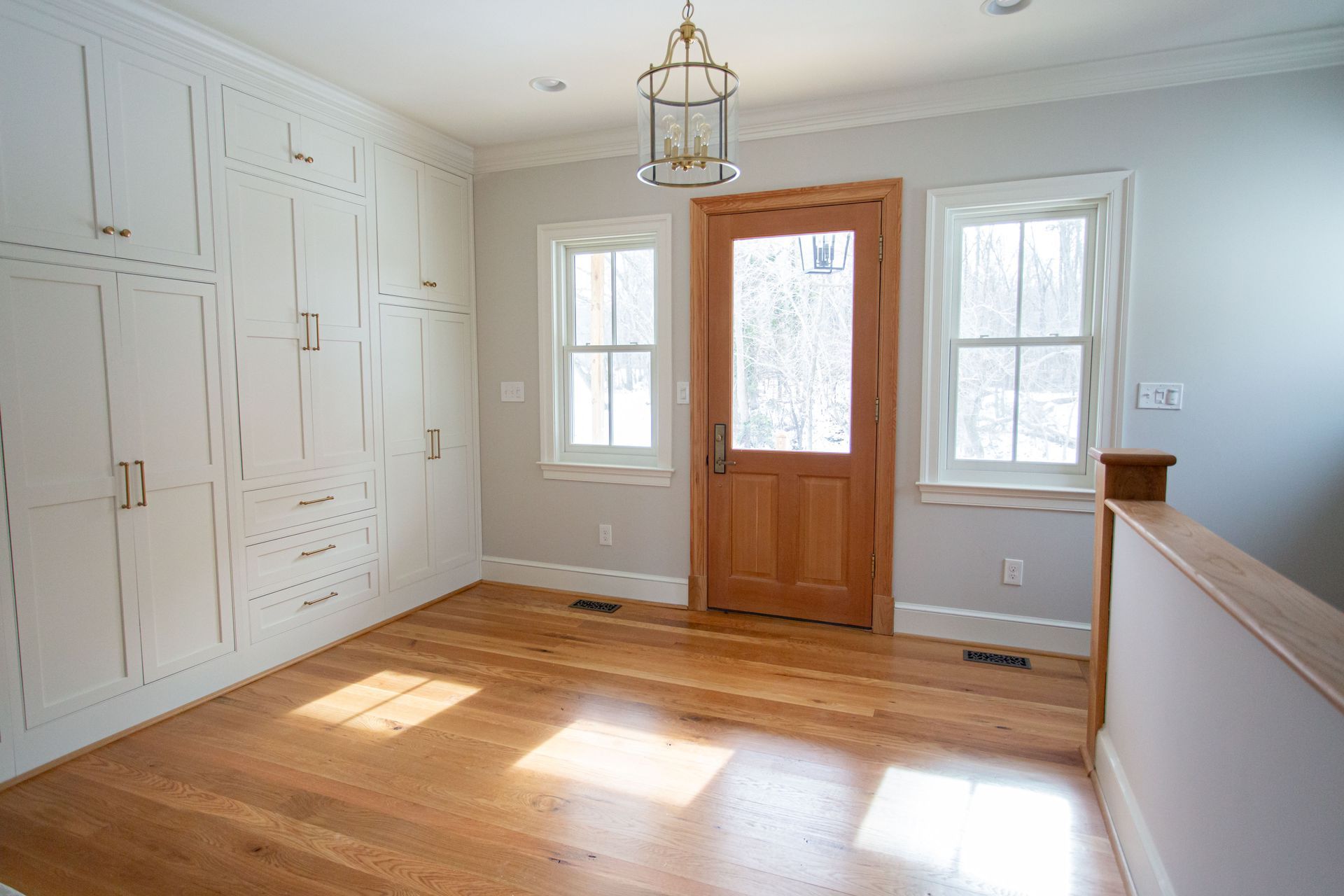 Room with wooden floor, white cabinets, brown door, two windows, and a hanging light fixture.