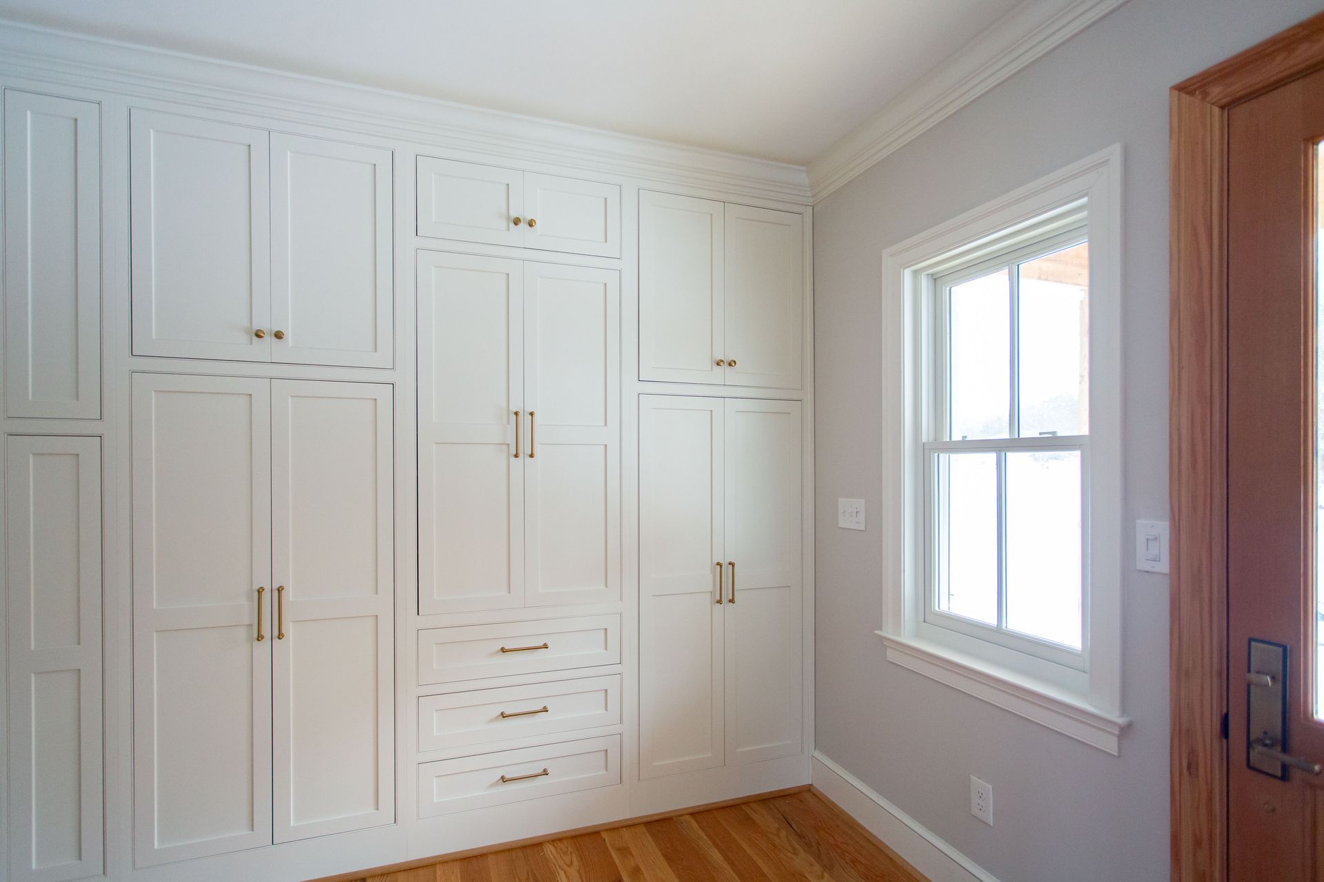 White built-in storage cabinets with drawers, next to a window and wooden door, in a room with hardwood floors.