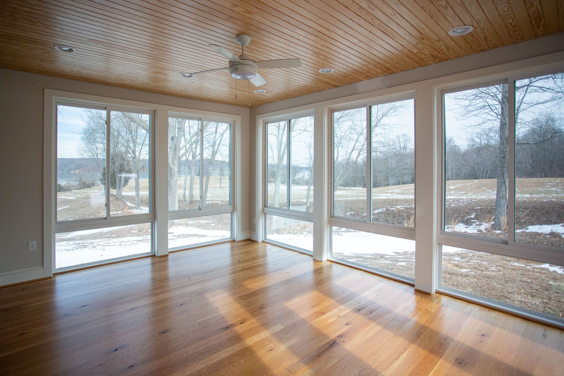 Sunroom with wooden floors, large windows overlooking a snowy field and trees, and a wood-paneled ceiling.