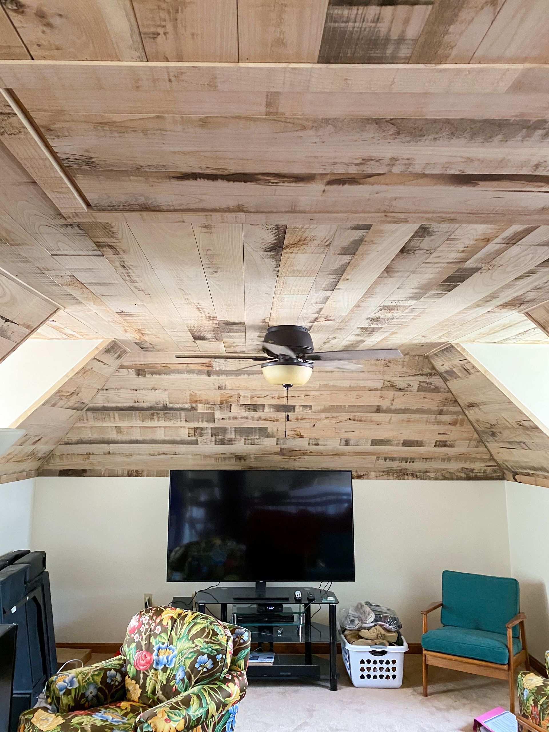 Living room with wood-paneled ceiling and TV centered on a white wall. Furniture includes a floral armchair and teal chair.