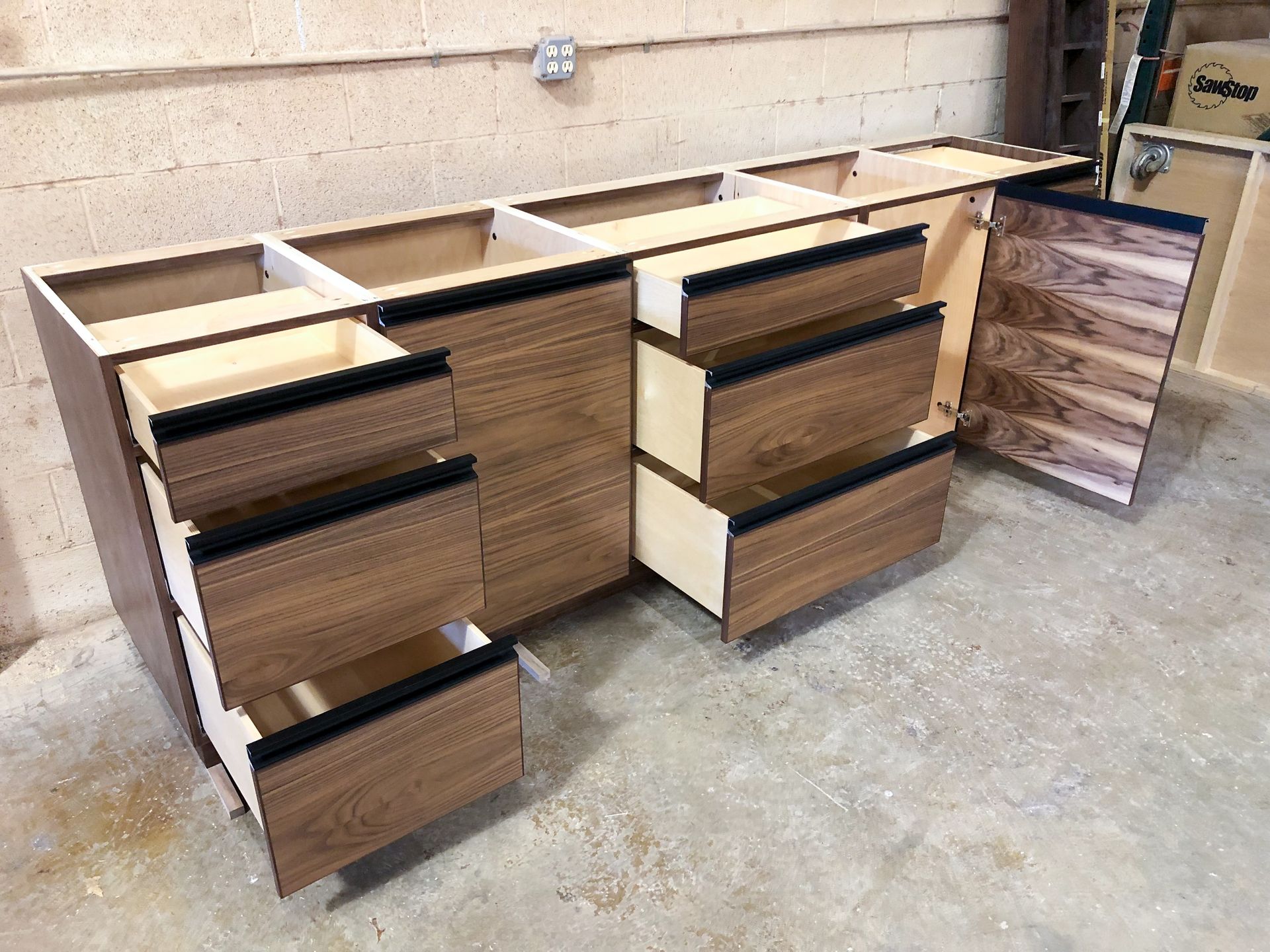 Wooden cabinet construction in progress; drawers and doors partially installed. Walnut grain finish, black trim.