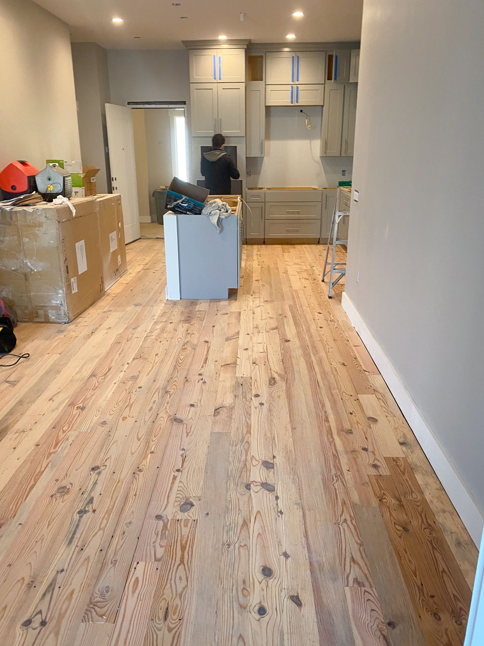 Wooden floor being installed in a kitchen area. Cabinets in the background, a person works.