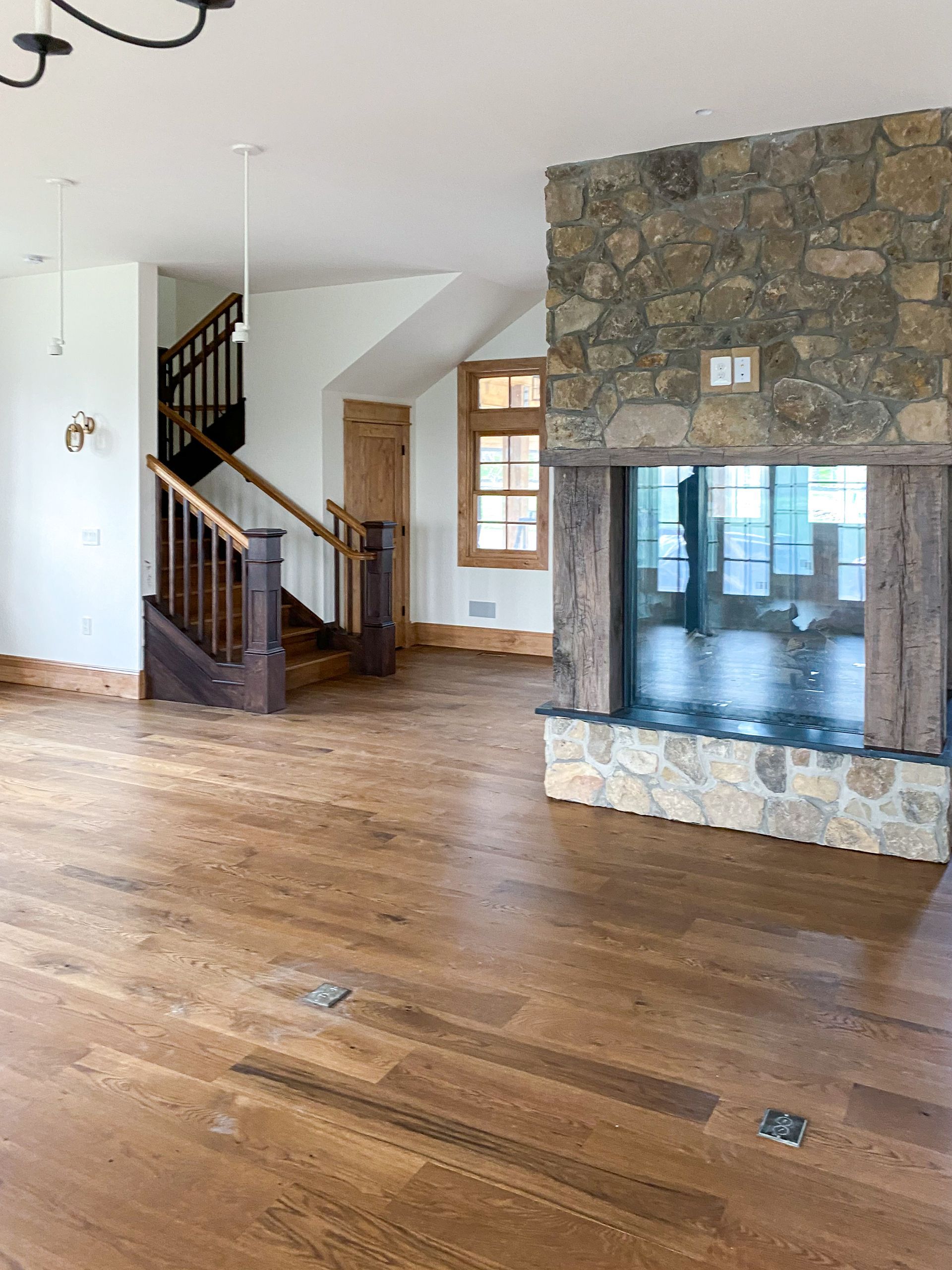 Spacious room with wood floors, stone fireplace, and a staircase. Sunlight enters from the window.