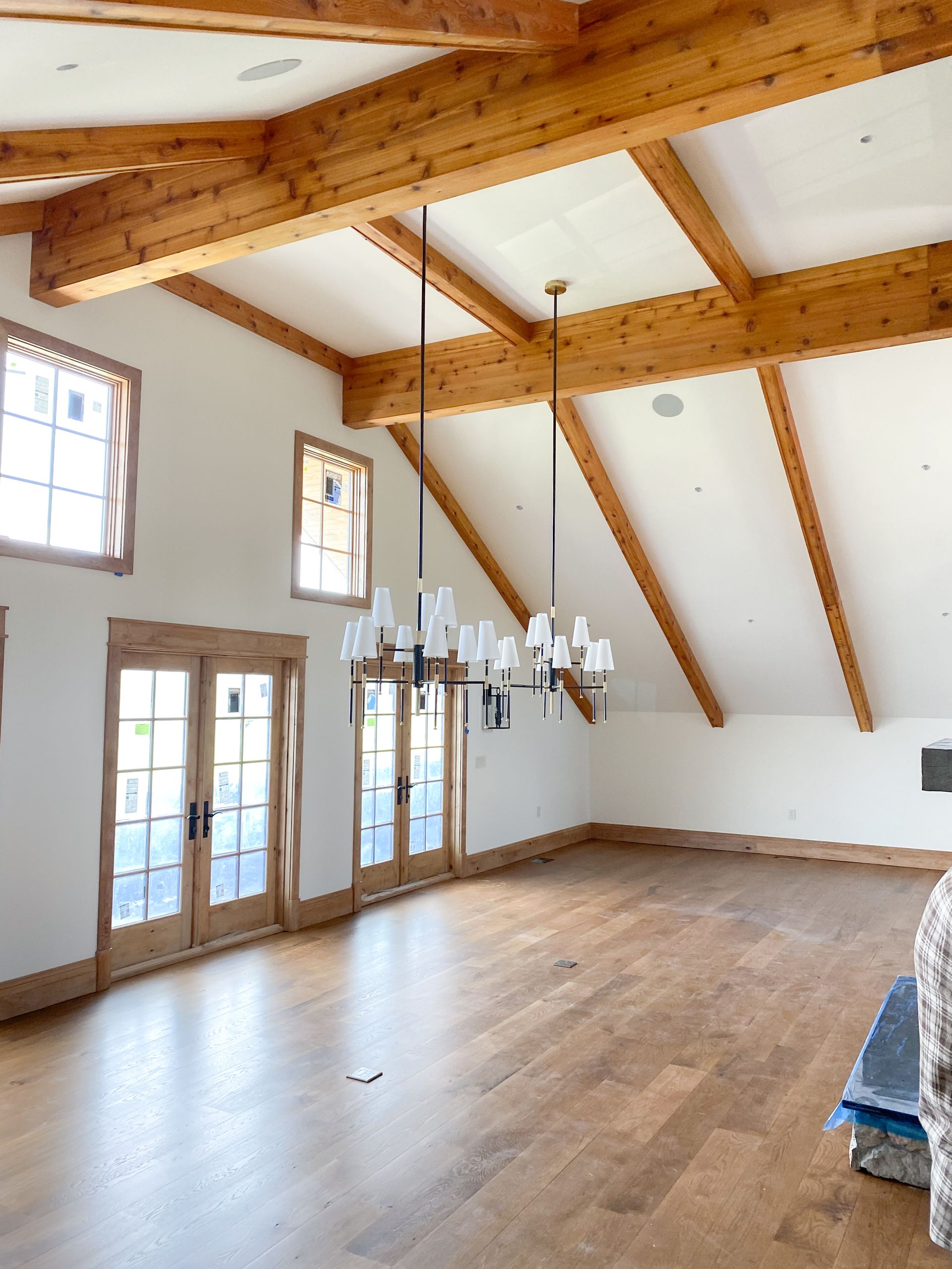 Spacious room with wood beams, light-colored walls, and hardwood floors. Two chandeliers hang from the ceiling.