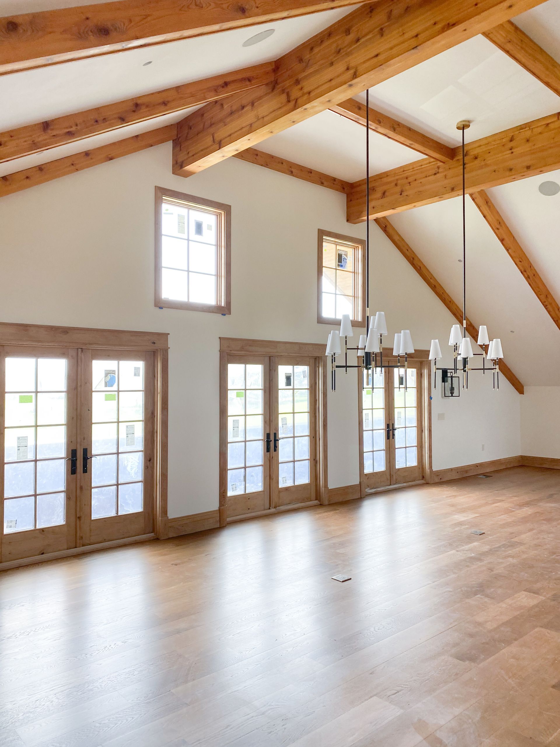 Spacious room with wood beams, light wood floors, and French doors. Modern chandelier hangs.