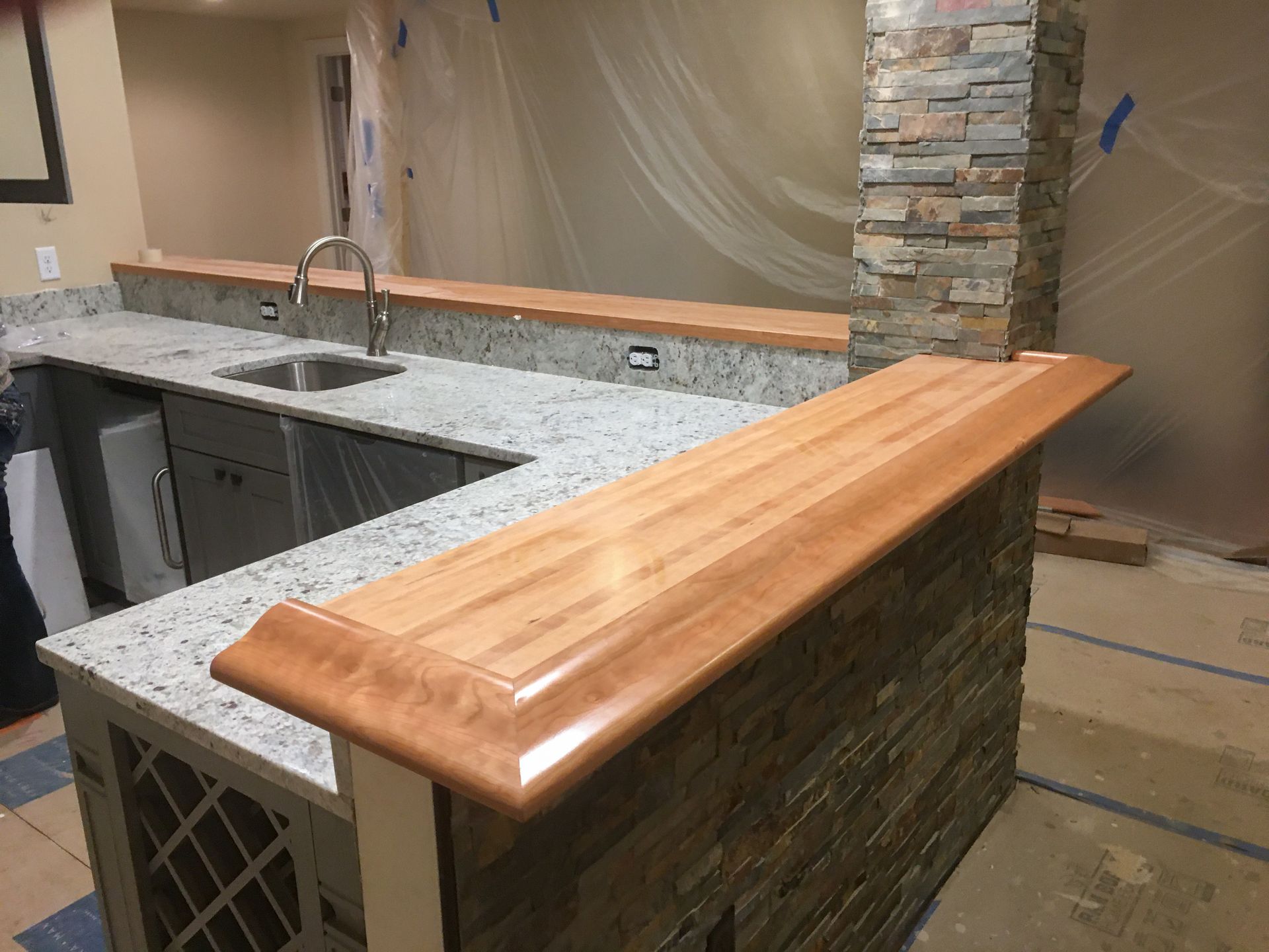 L-shaped bar with stone and wood accents. Granite countertop, stainless steel sink and appliances. Light wood trim.