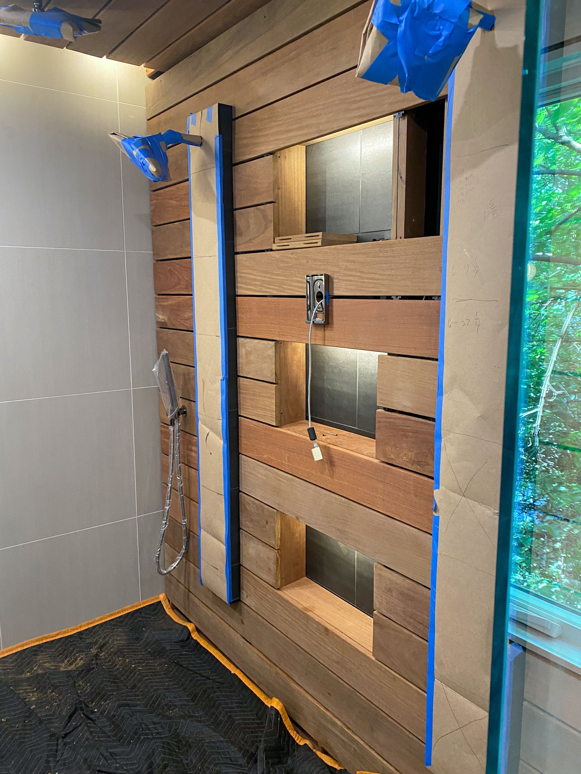 Wooden shower wall with open cubby holes, taped with blue tape and paper. Window visible.