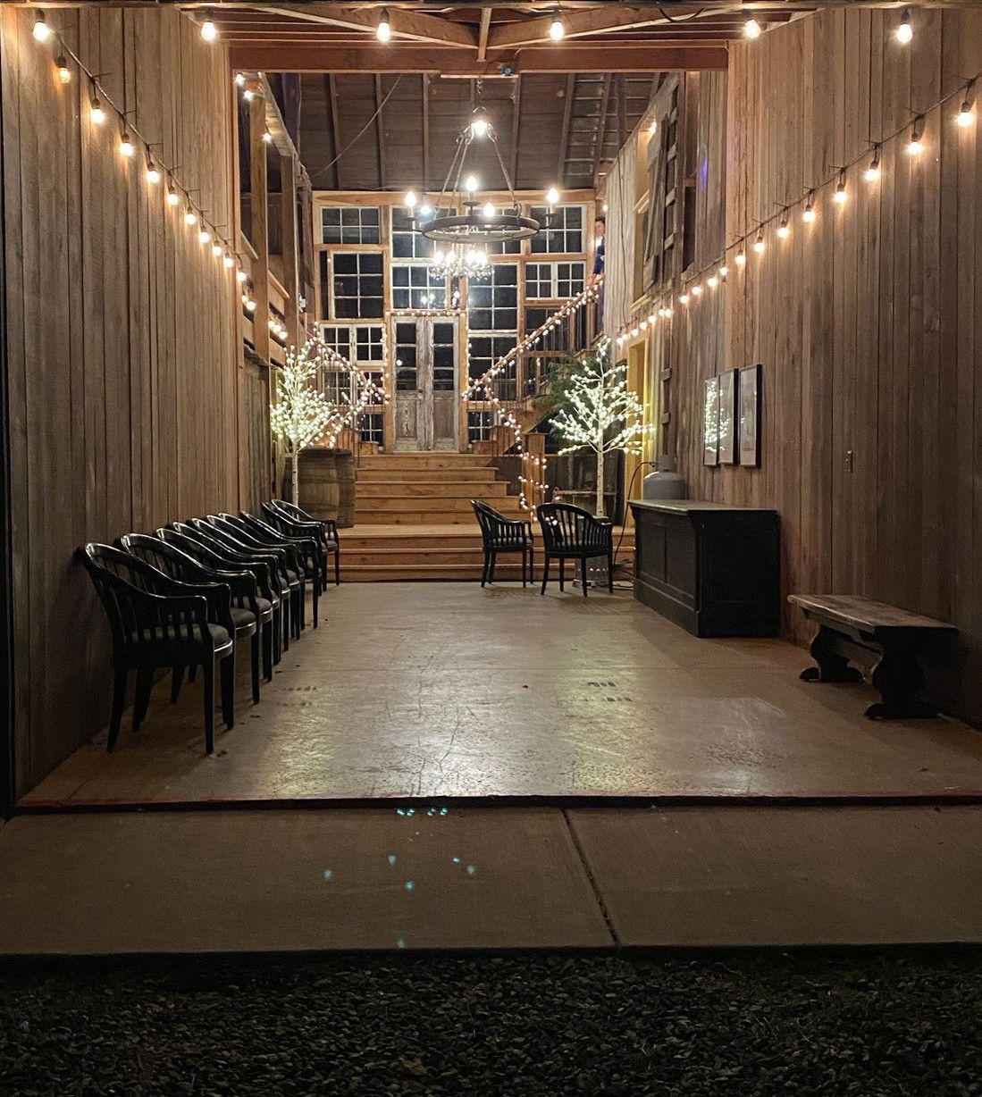 Barn interior with rows of chairs, stairs, and string lights. Wooden walls and a dark bar.