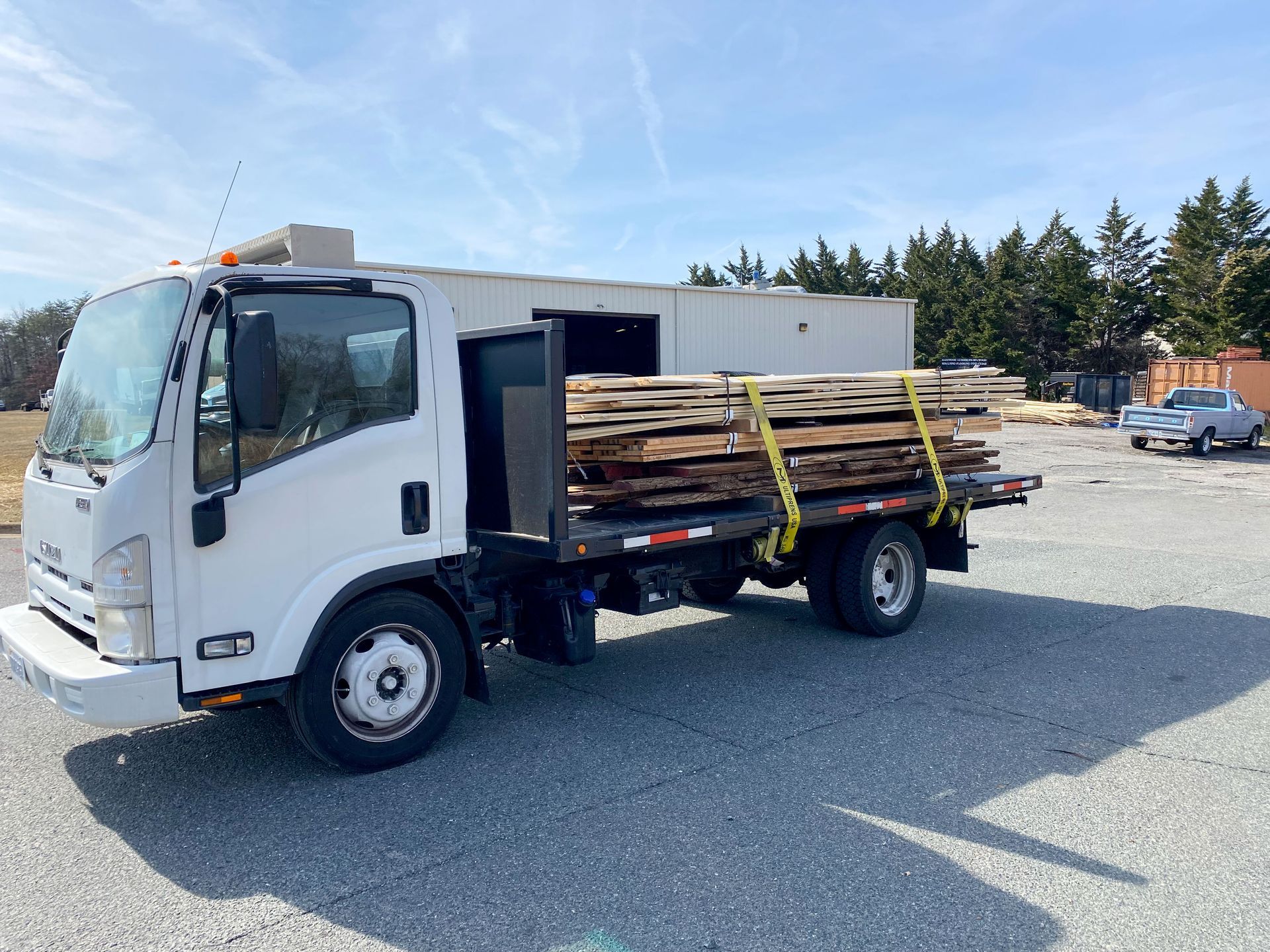 White truck loaded with wood planks on a gravel lot under a blue sky.