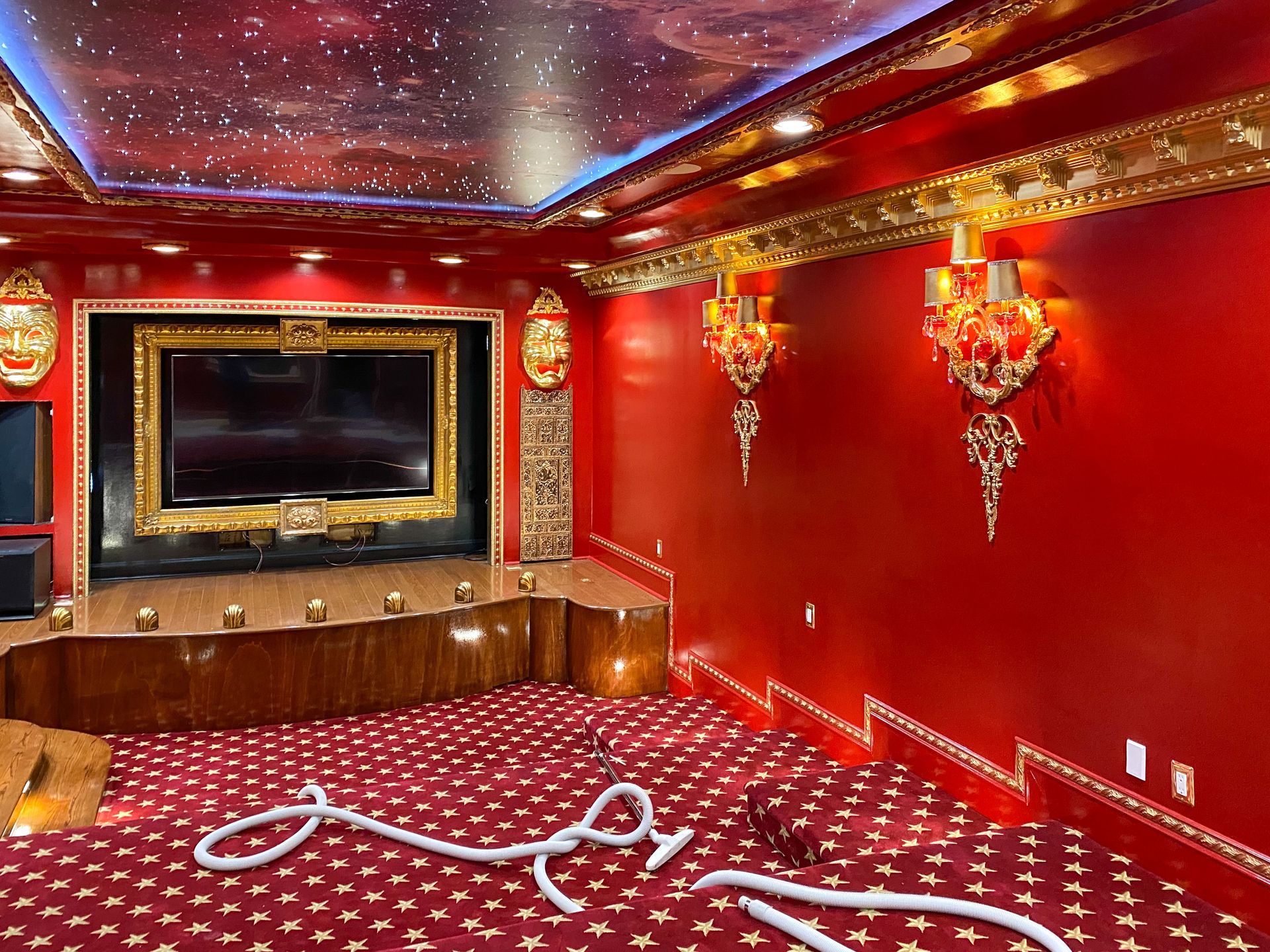 Red and gold home theater room with galaxy ceiling, large TV, ornate sconces, and red carpet.