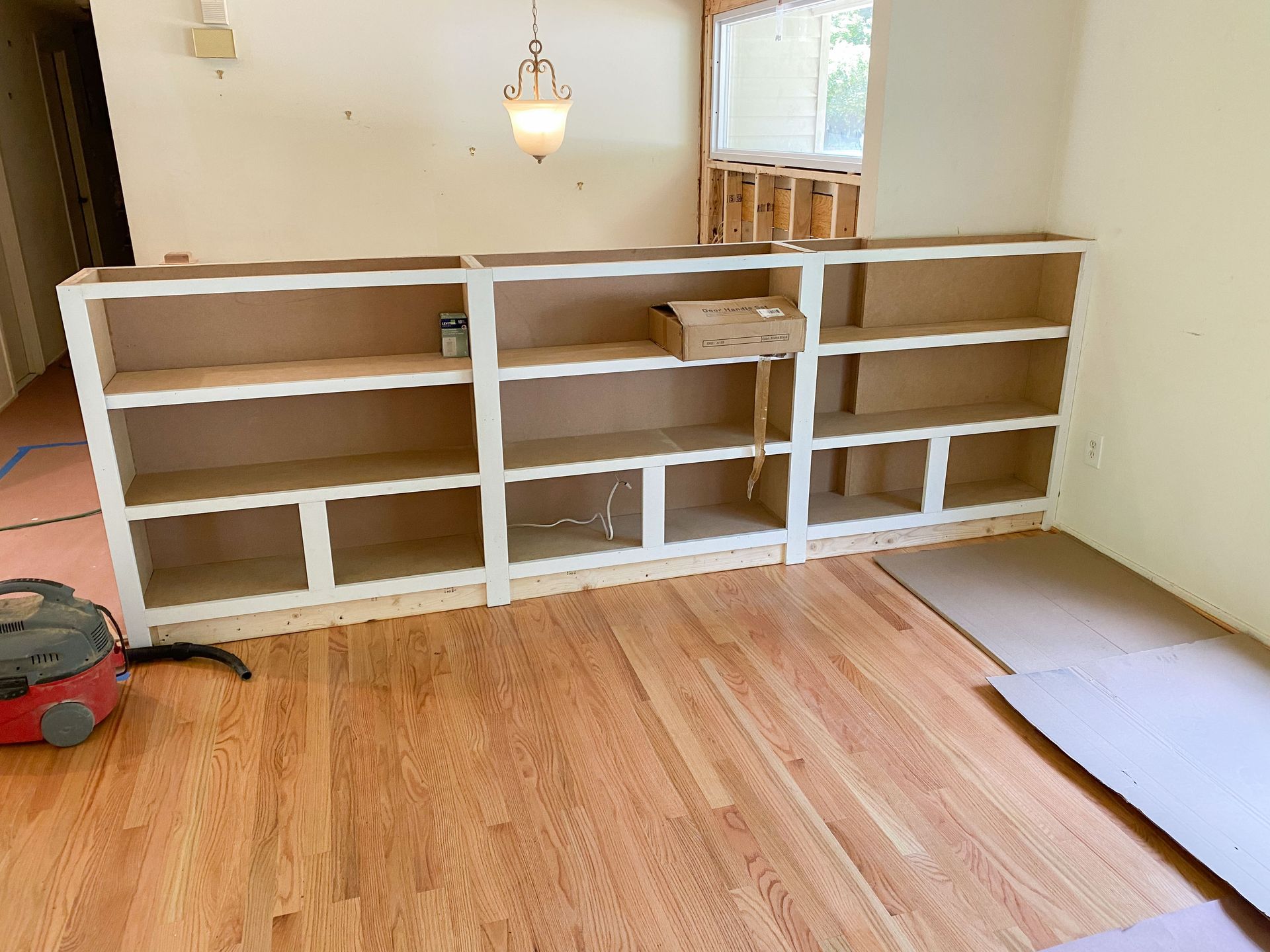 White built-in bookshelves against a light wall, over wooden floors; a vacuum cleaner sits nearby.