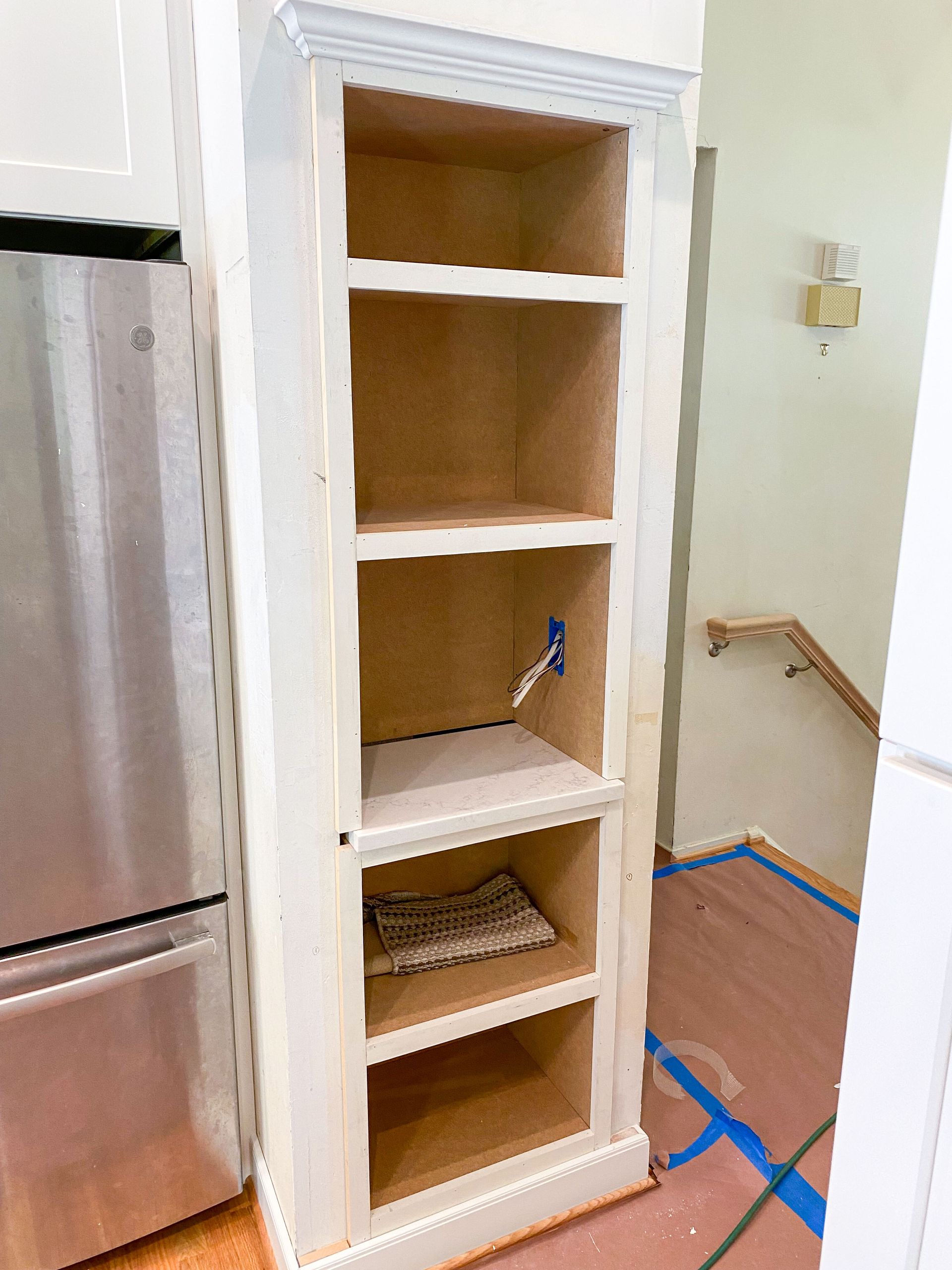 A tall, narrow, unfinished built-in cabinet with shelves in a kitchen, next to a refrigerator.
