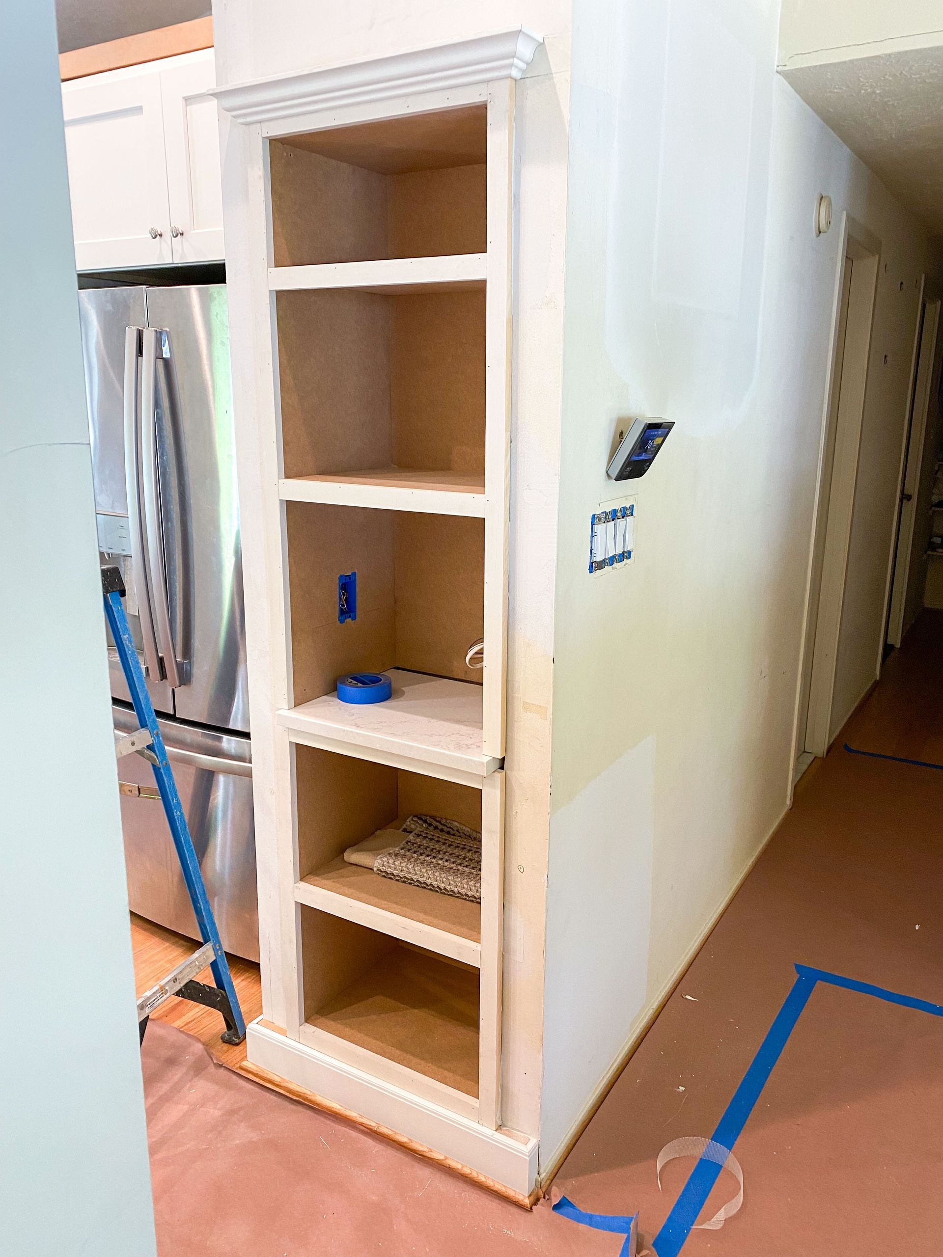 Wooden built-in shelving unit, unfinished, next to a hallway. Blue tape on the floor.