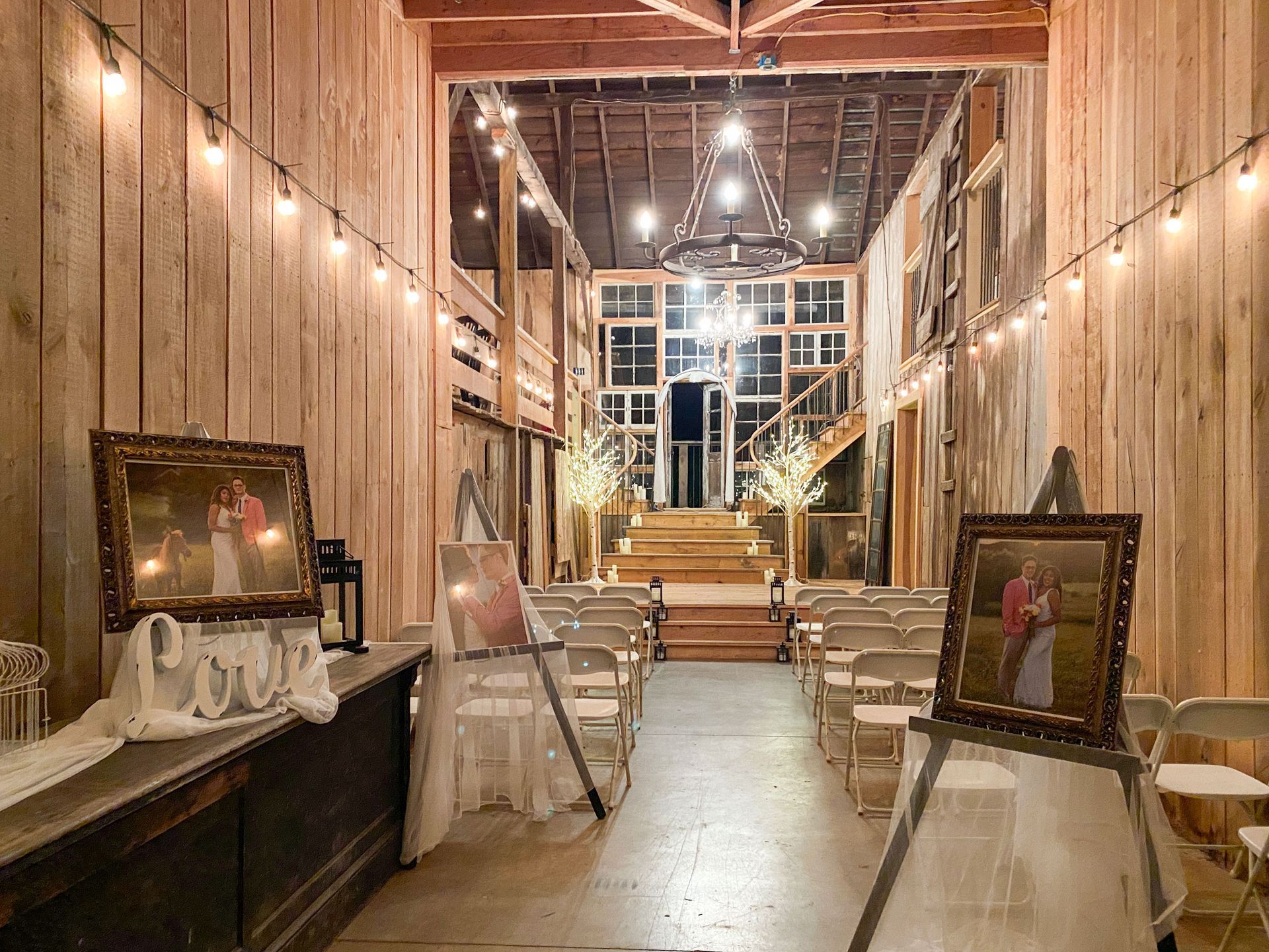 Inside a rustic barn wedding venue, rows of chairs face a raised altar. Portraits flank the aisle, lit by string lights.