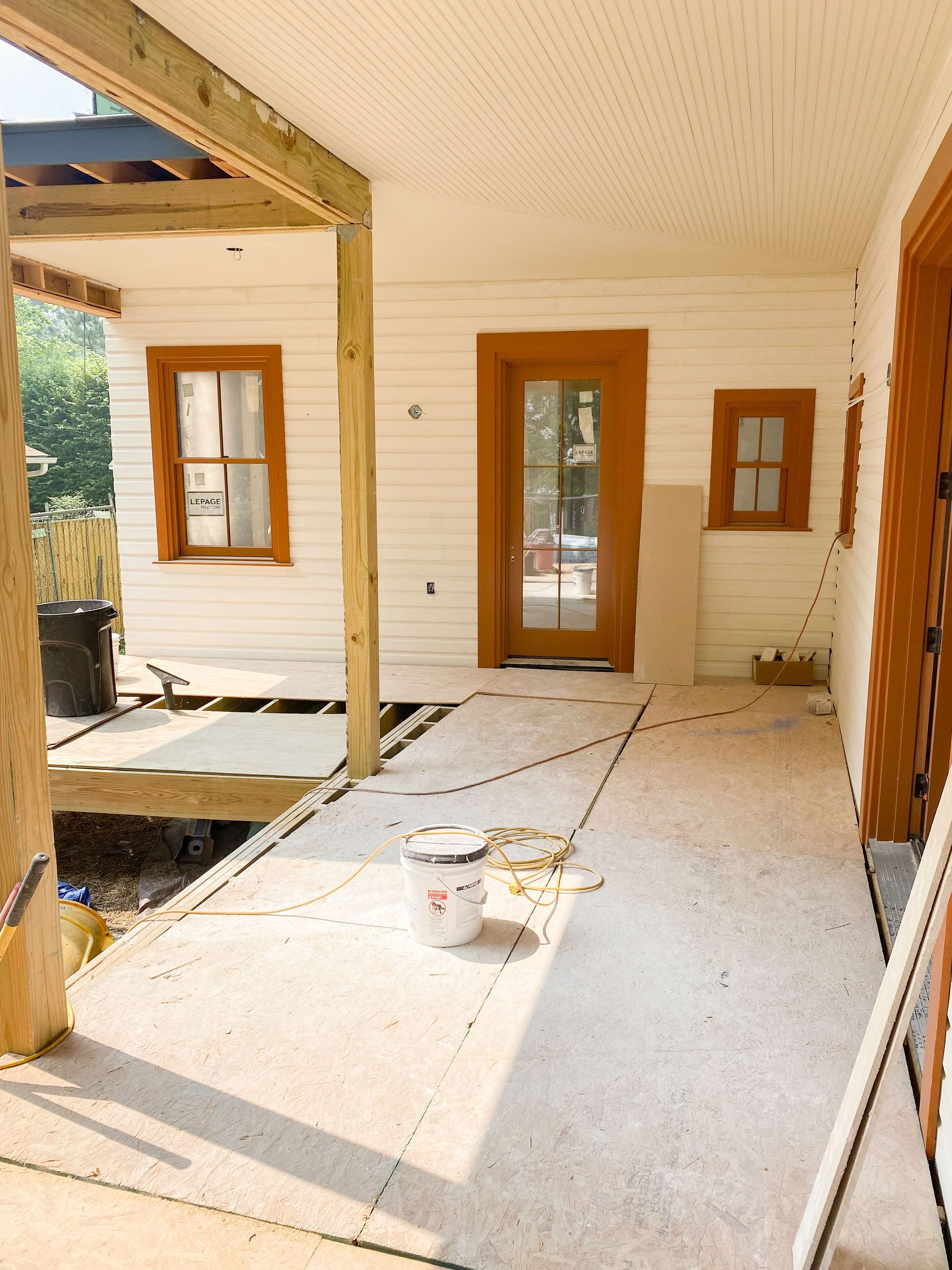 Porch under construction with white siding, wood trim, and a door, with exposed beams overhead.