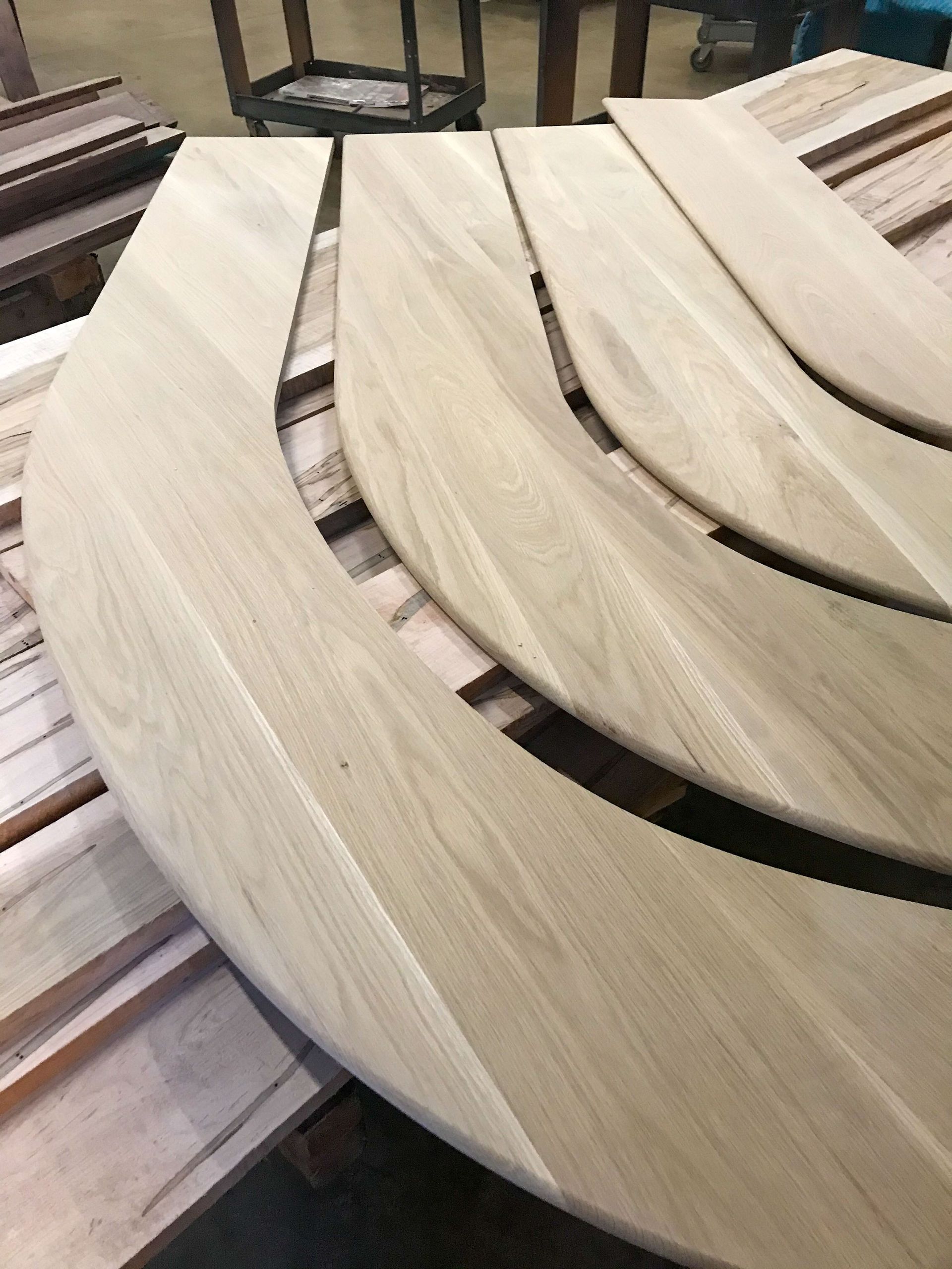 Curved wooden table sections stacked, ready for assembly. Light wood tone. Industrial setting.