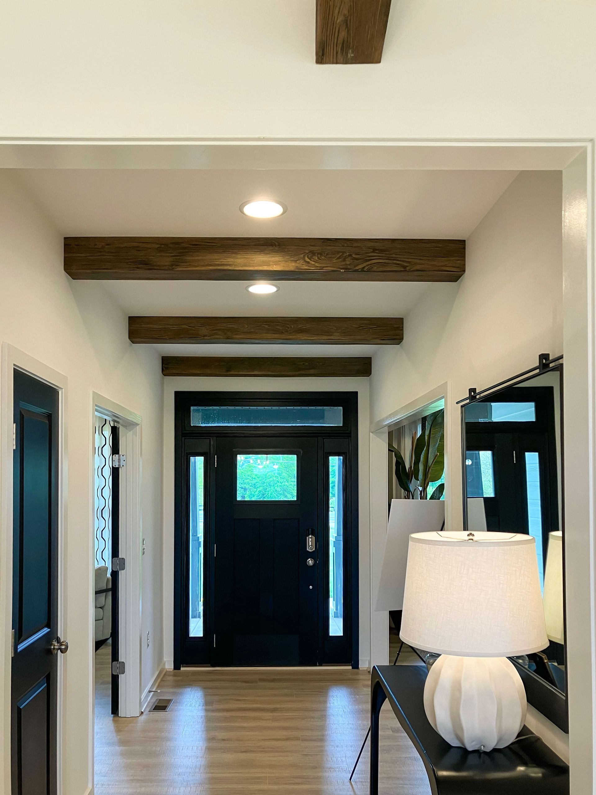 Hallway with dark door, wood beams, recessed lights, and a table with a lamp.