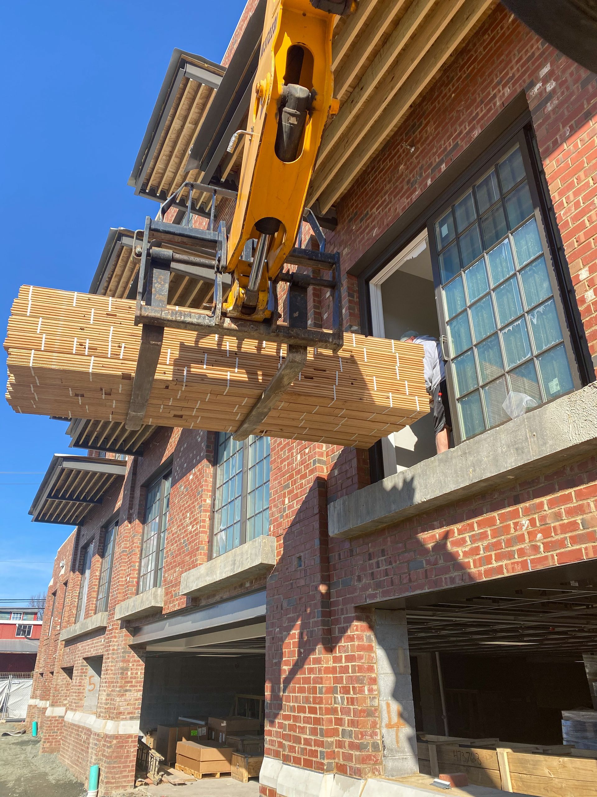 A forklift lifting a pallet of wood toward the window of a brick building under construction.