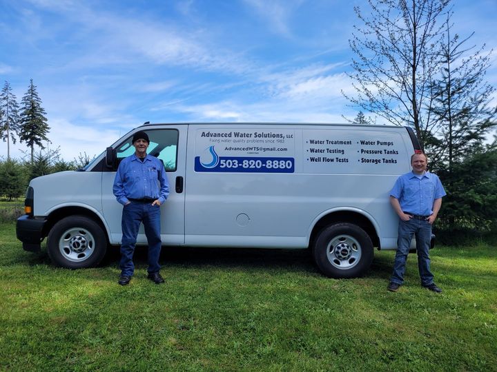 Two workers stand beside a white service van on a grassy field under a blue sky.