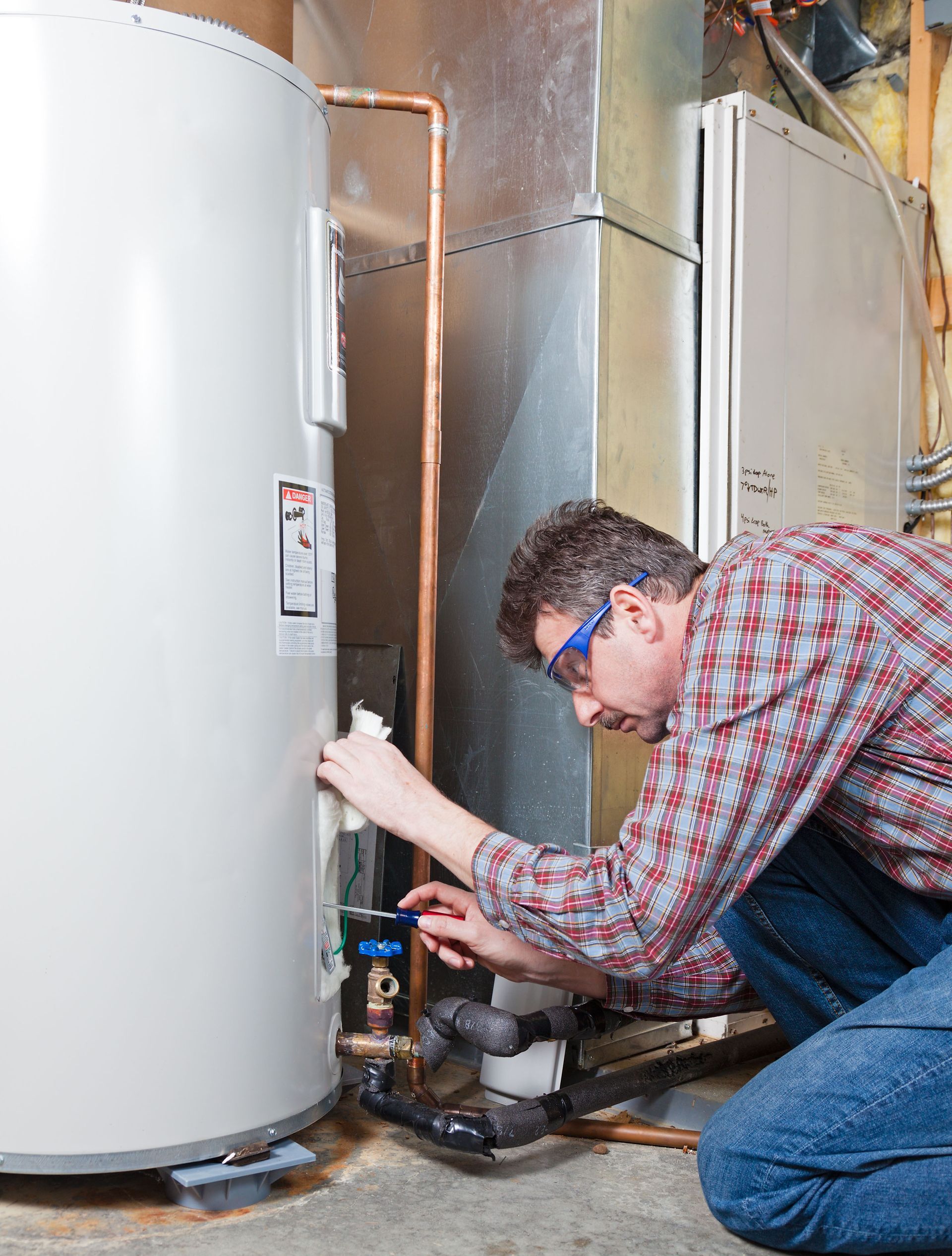 Technician kneeling by a water heater, inspecting pipes and fittings in a utility room.