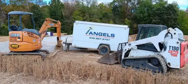 Two construction machines beside a white ANGELS truck in a brushy field