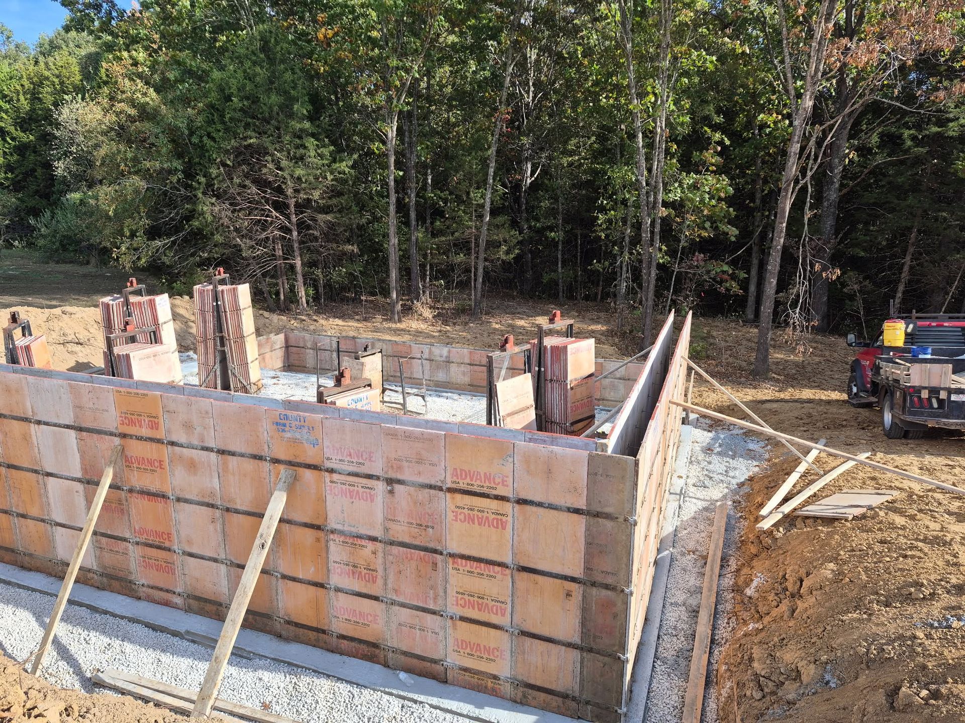 Partially built concrete foundation wall at a wooded construction site, with a pickup truck nearby