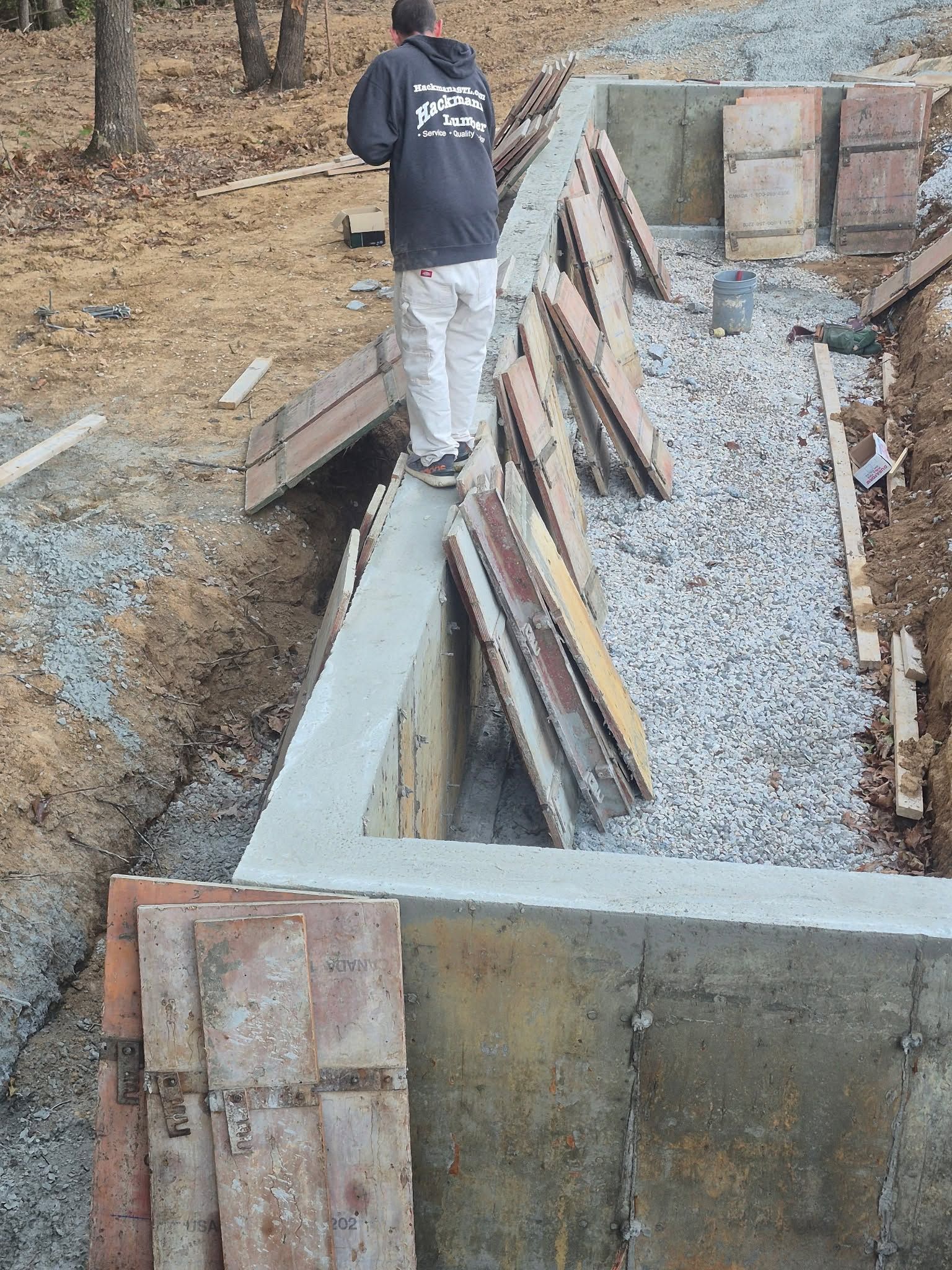 Worker standing on cinder blocks beside a concrete foundation trench with wooden braces and gravel