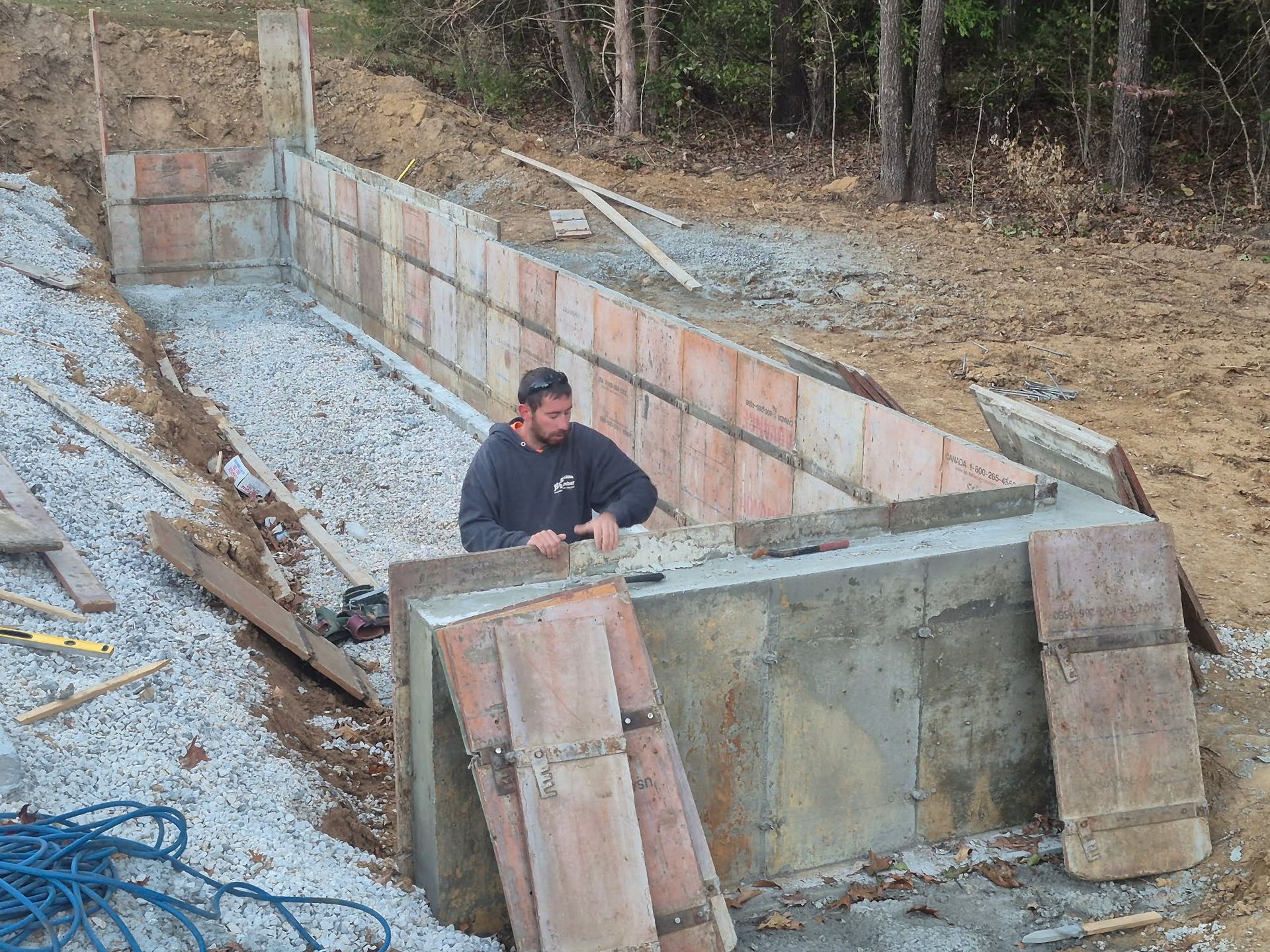 Worker beside a concrete retaining wall under construction on a sloped, wooded site.