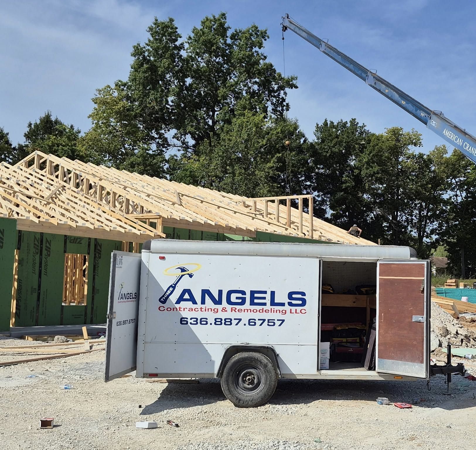 Construction site with a white ANGELS work van beside a house under roof framing, crane in background