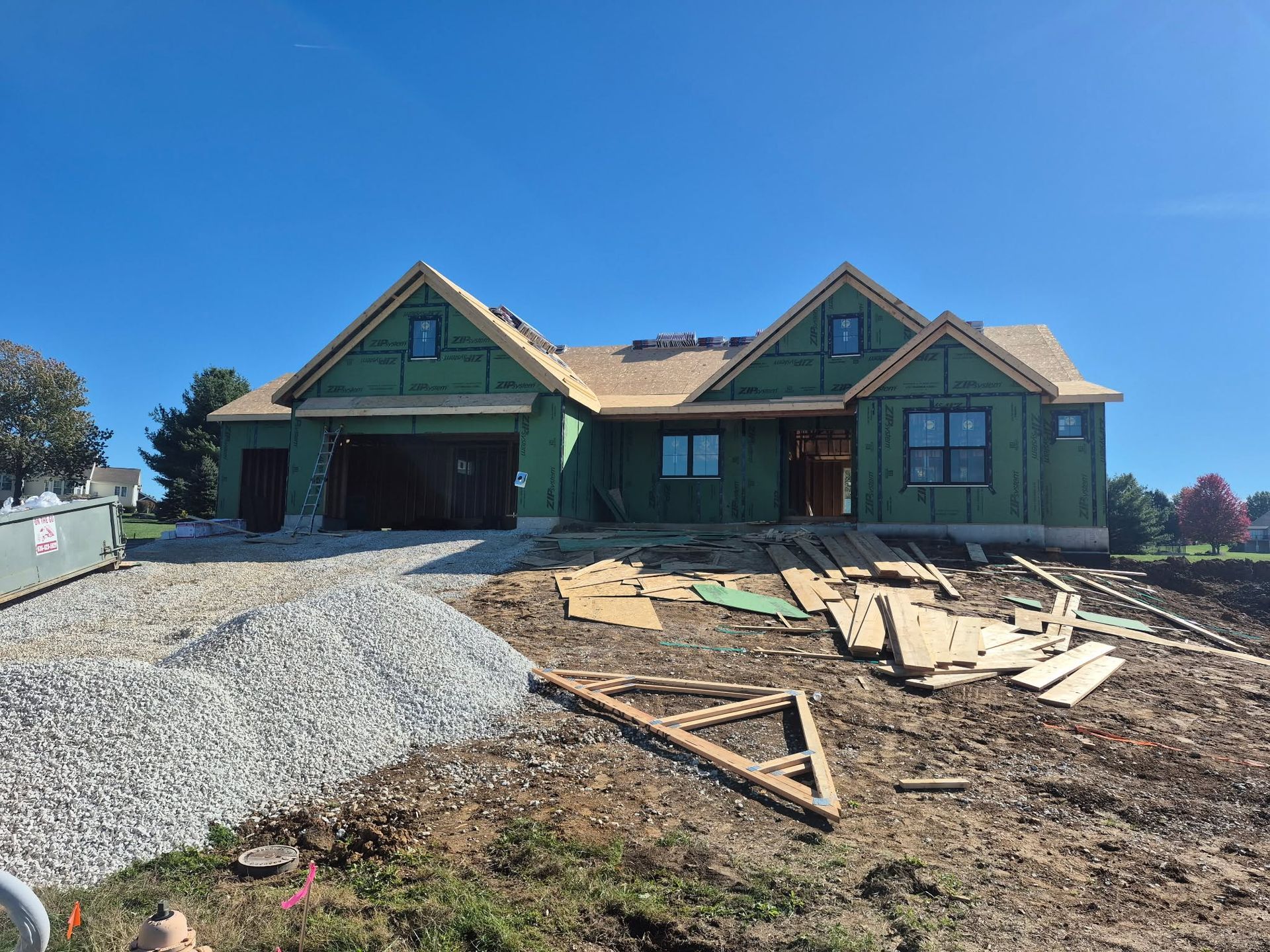 House under construction with exposed framing, gravel driveway, and lumber stacked in the yard under a blue sky