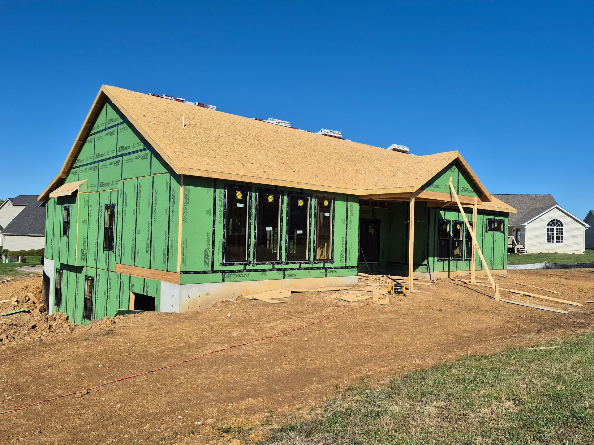 Green house under construction with tan roof sheathing on a dirt lot under a clear blue sky