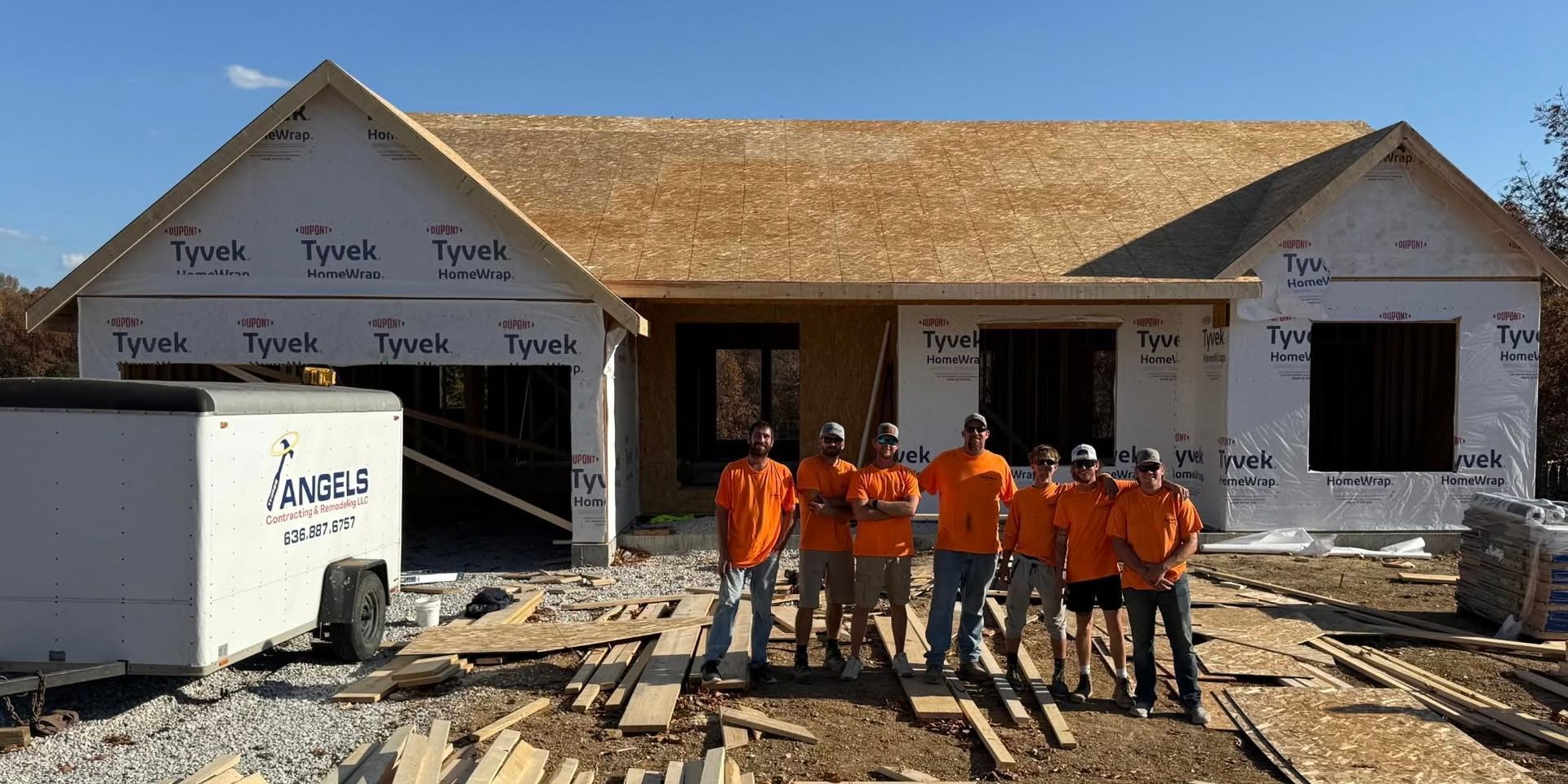 Construction crew in orange vests stands outside a house under construction with exposed framing.