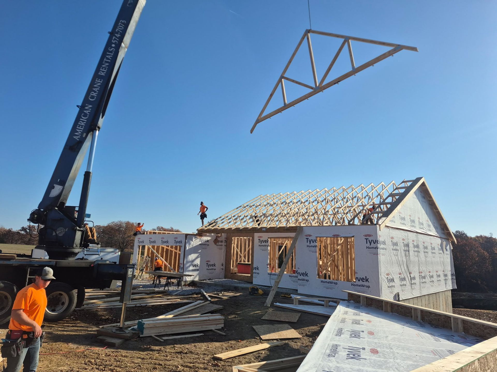 Workers and a crane assemble a house frame at a construction site, with roof trusses overhead.