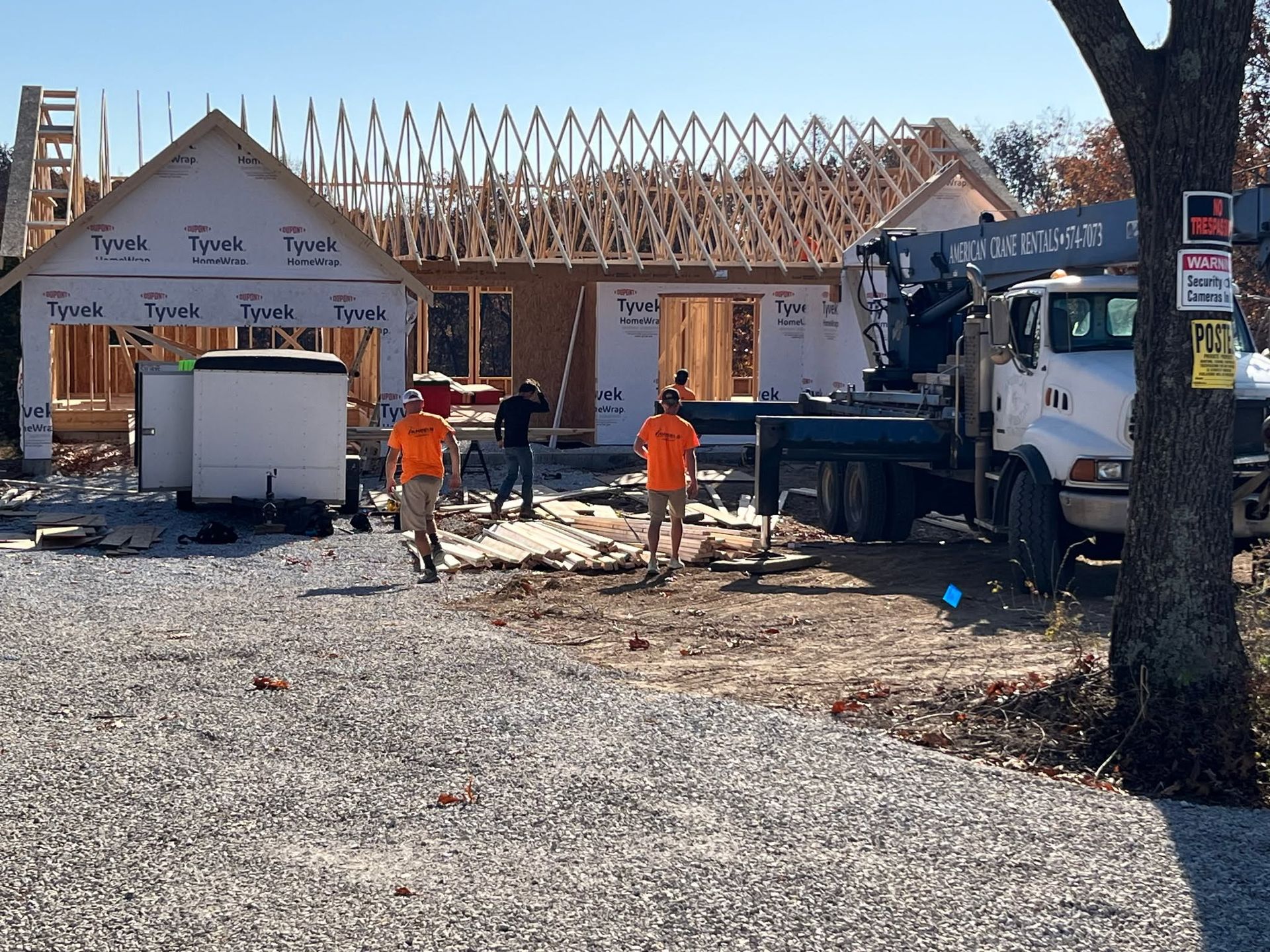 Construction workers building a house frame beside a white truck and piles of lumber on a gravel lot