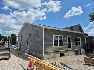 House exterior under construction with gray siding, ladders, and a partially finished yard under a blue sky