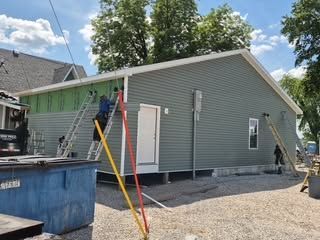 Workers painting gray house exterior with ladders and blue dumpster under a sunny sky