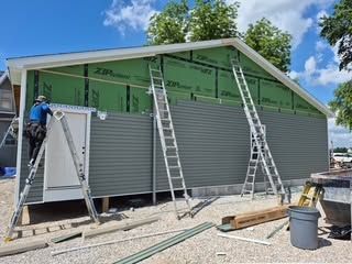 Workers installing gray siding on a house under construction, with ladders and green sheathing visible.