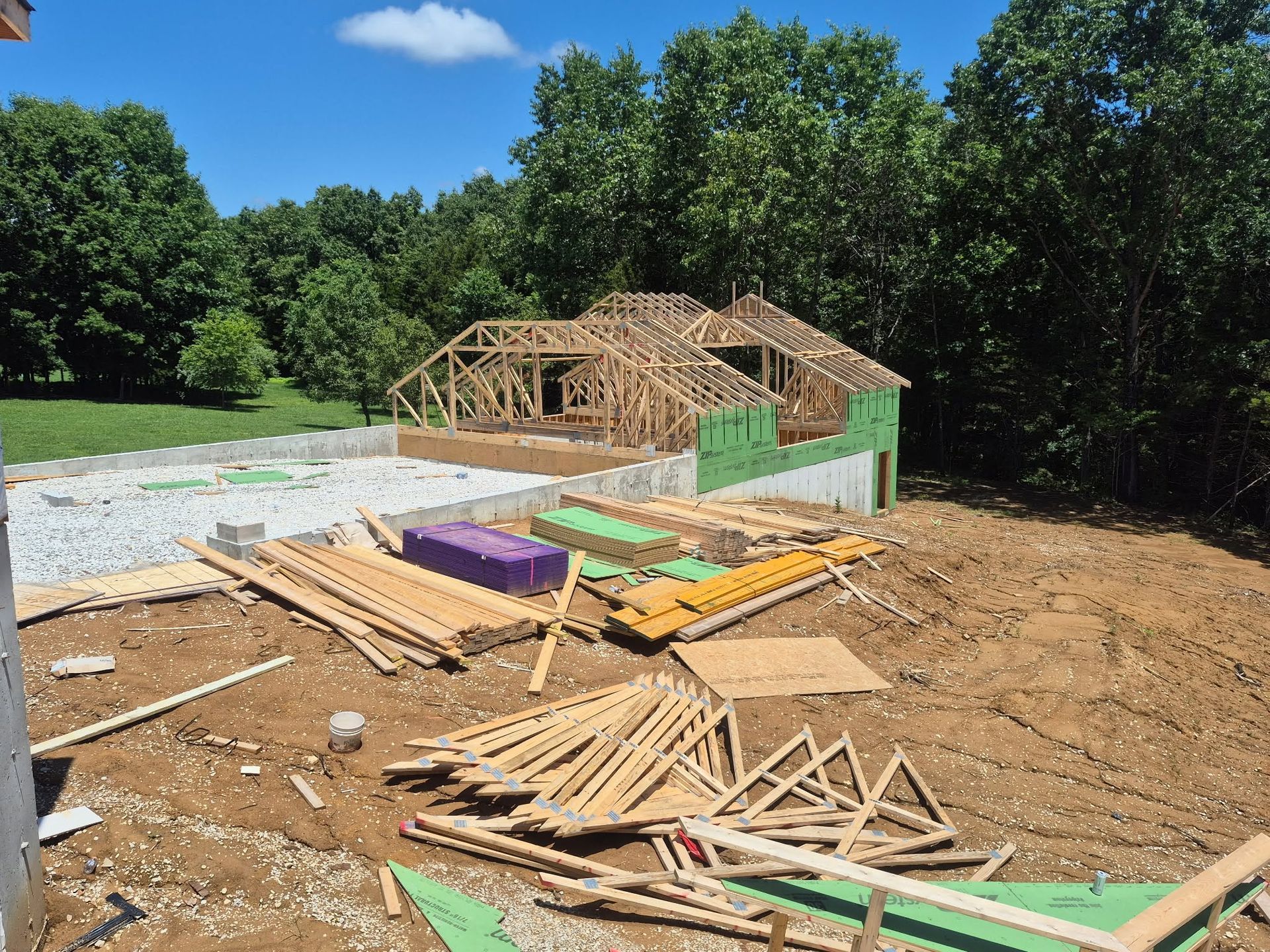 Partially built house frame on a cleared lot with lumber scattered in the foreground and trees behind