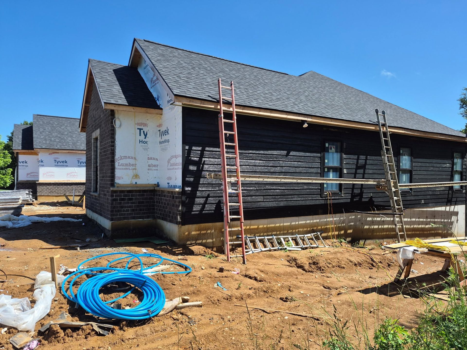 Wooden house under construction with ladders, scaffolding, and blue hose on a dirt lot