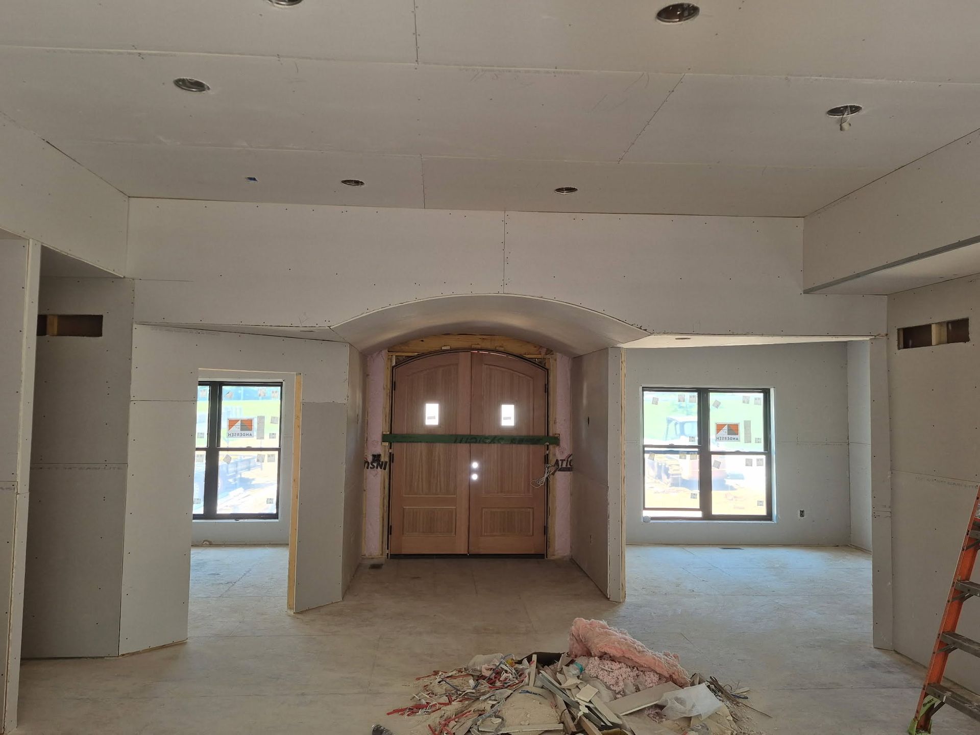 Empty unfinished room with arched wooden double doors, two windows, and construction debris on the floor
