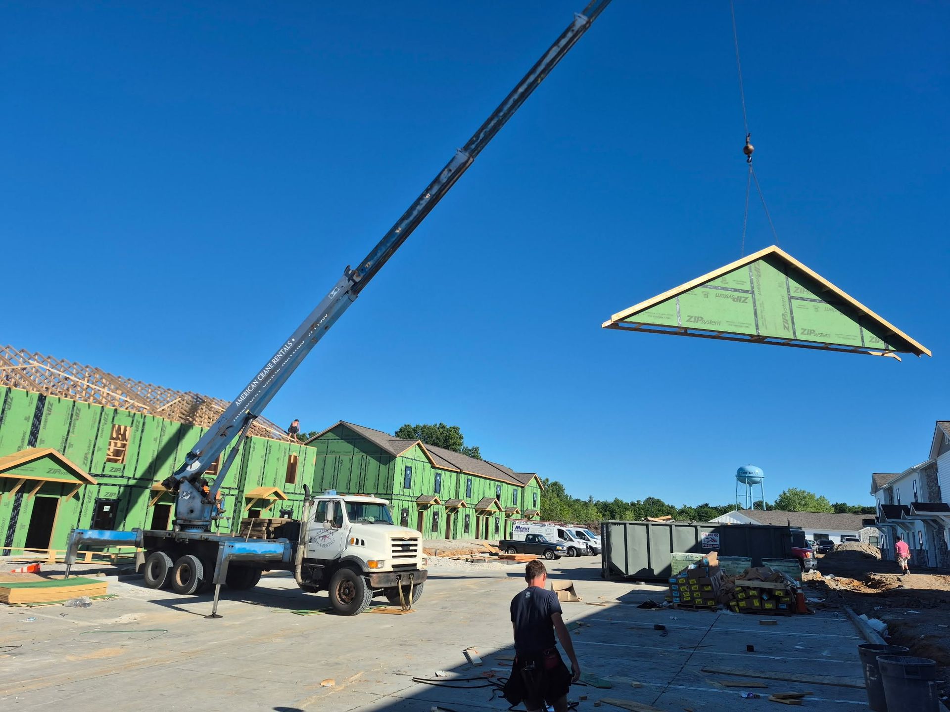 Crane lifting a green roof panel at a construction site with workers and buildings in the background
